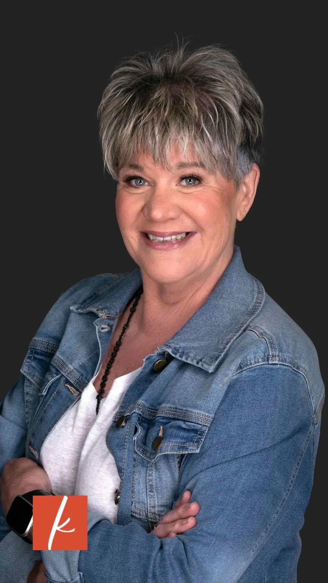 Smiling woman with short gray hair and crossed arms, wearing a denim jacket and white top against a dark studio backdrop.