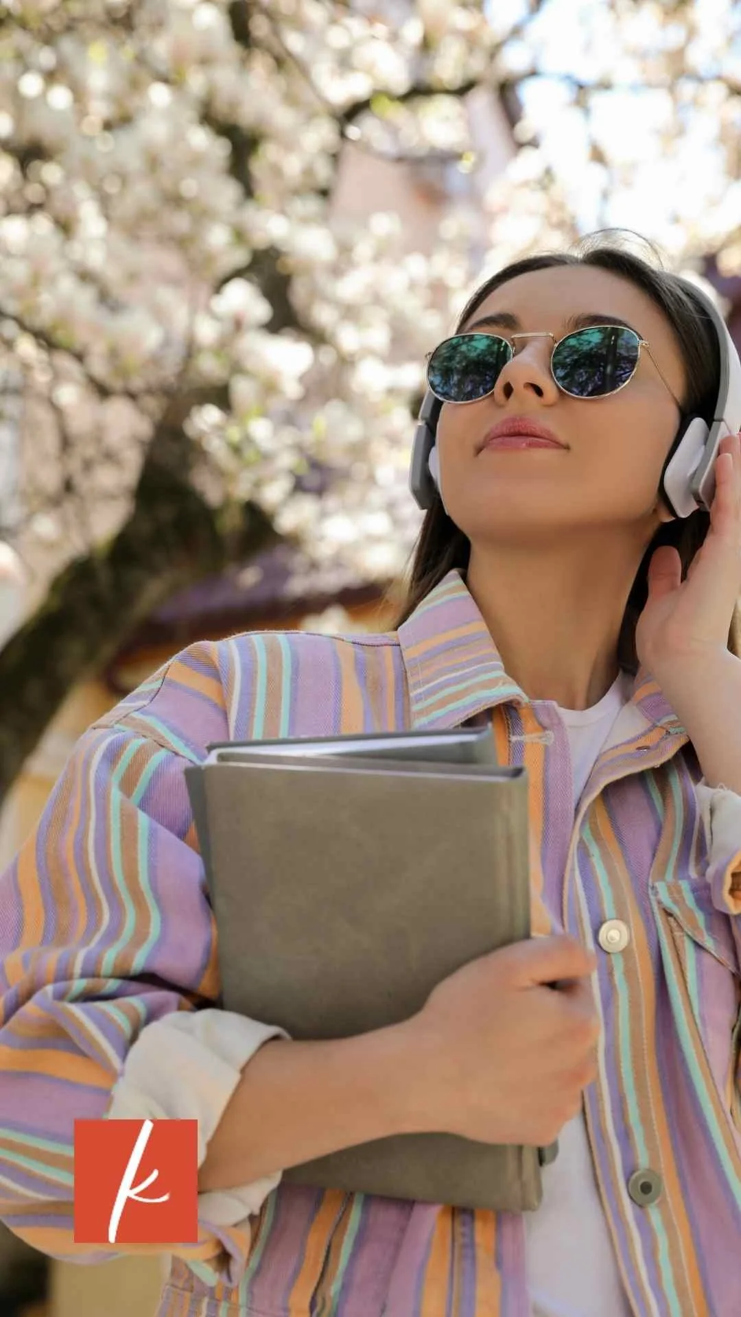 Person wearing headphones and sunglasses holding a notebook while listening outdoors among blooming trees.