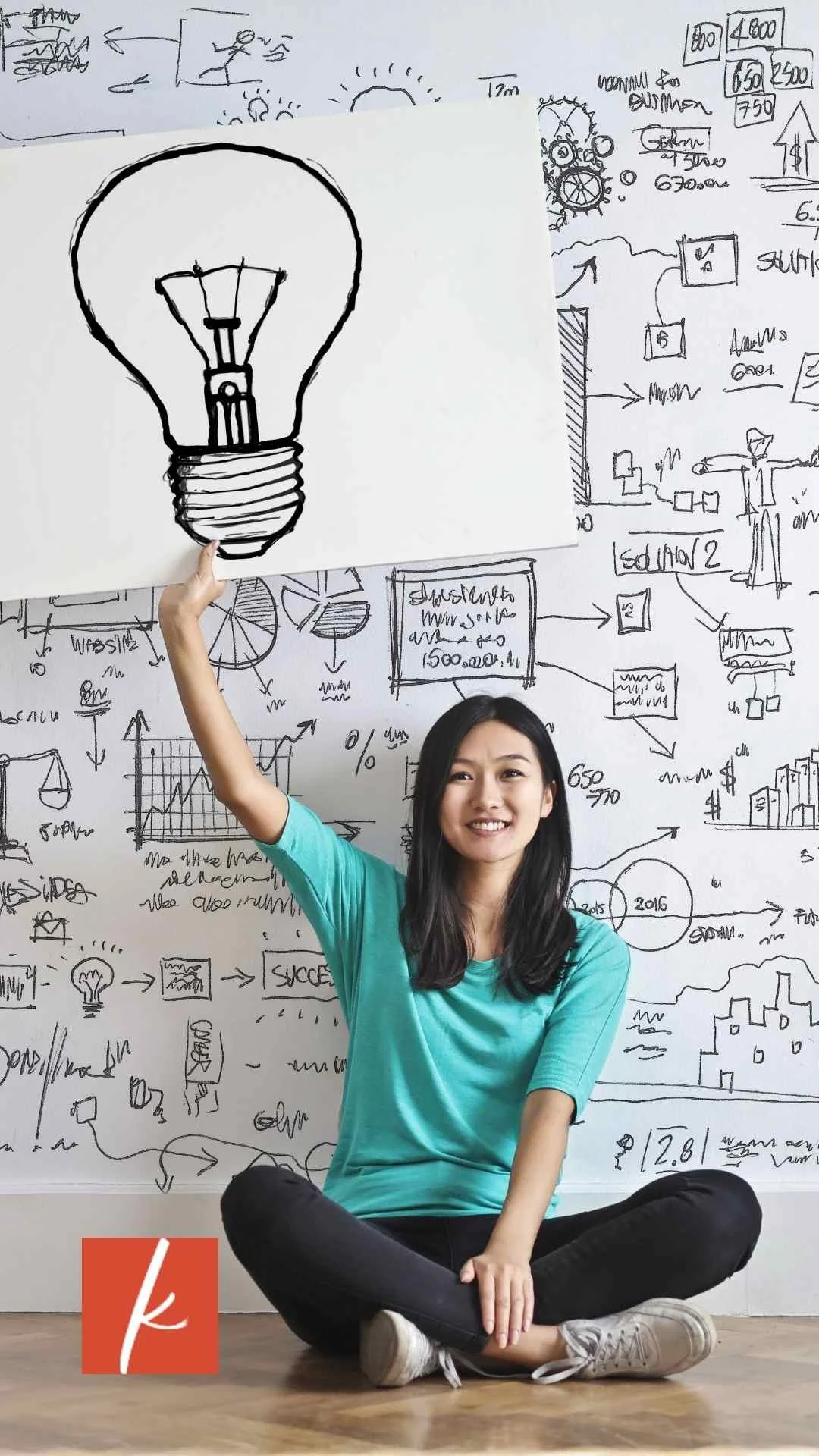 Woman sitting cross-legged holding a drawing of a light bulb in front of a wall covered with planning sketches.