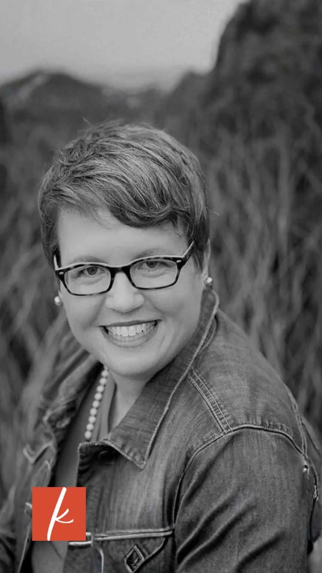 Black-and-white portrait of a smiling woman with short hair and glasses wearing a denim jacket outdoors.