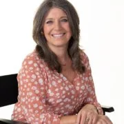 A woman with brown curly hair, wearing a salmon colour polka dot top, sitting on a chair, smiling, with a plain white background.