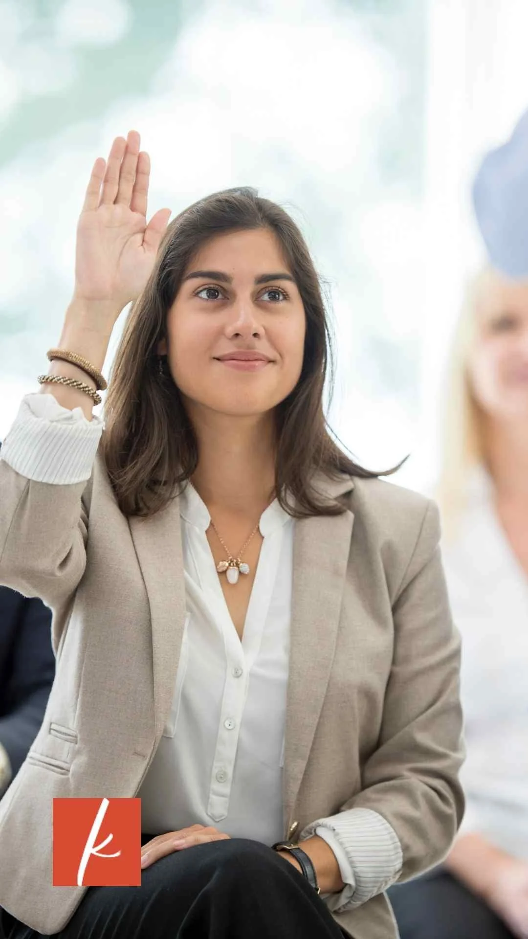 Woman wearing business casual attire sitting in a meeting raising her hand.