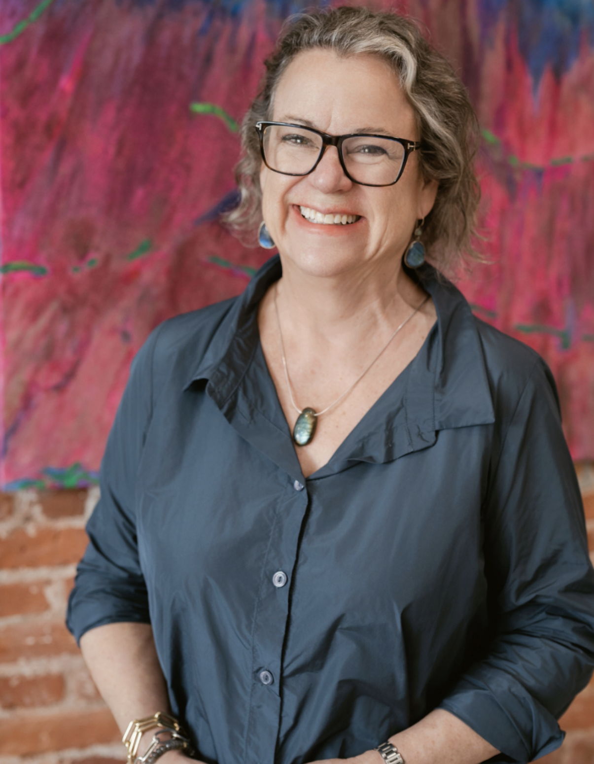 Portrait of a smiling woman with short, curly brown hair, wearing glasses and a blue top, against a pink and colourfulbackground.