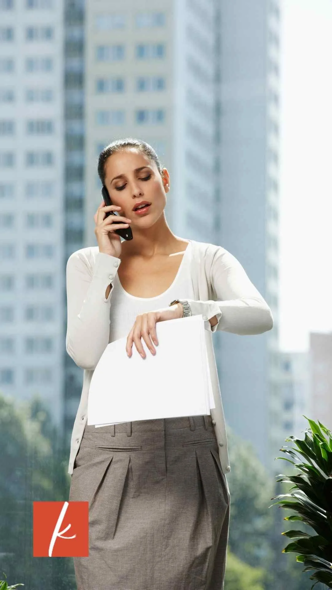 Woman on a phone call holding documents and checking the time, standing near a window with city buildings in the background.