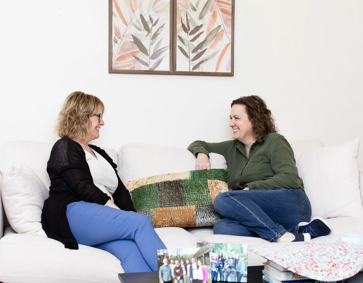 Two women sitting on a white sofa, smiling and talking to each other. One woman has blond hair and glasses, wearing a black cardigan and blue pants. The other has curly brown hair, wearing a green shirt and jeans. There is a colorful patchwork pillow between them, and a table with photographs and a gift bag in front of them. A framed abstract botanical artwork hangs above them.