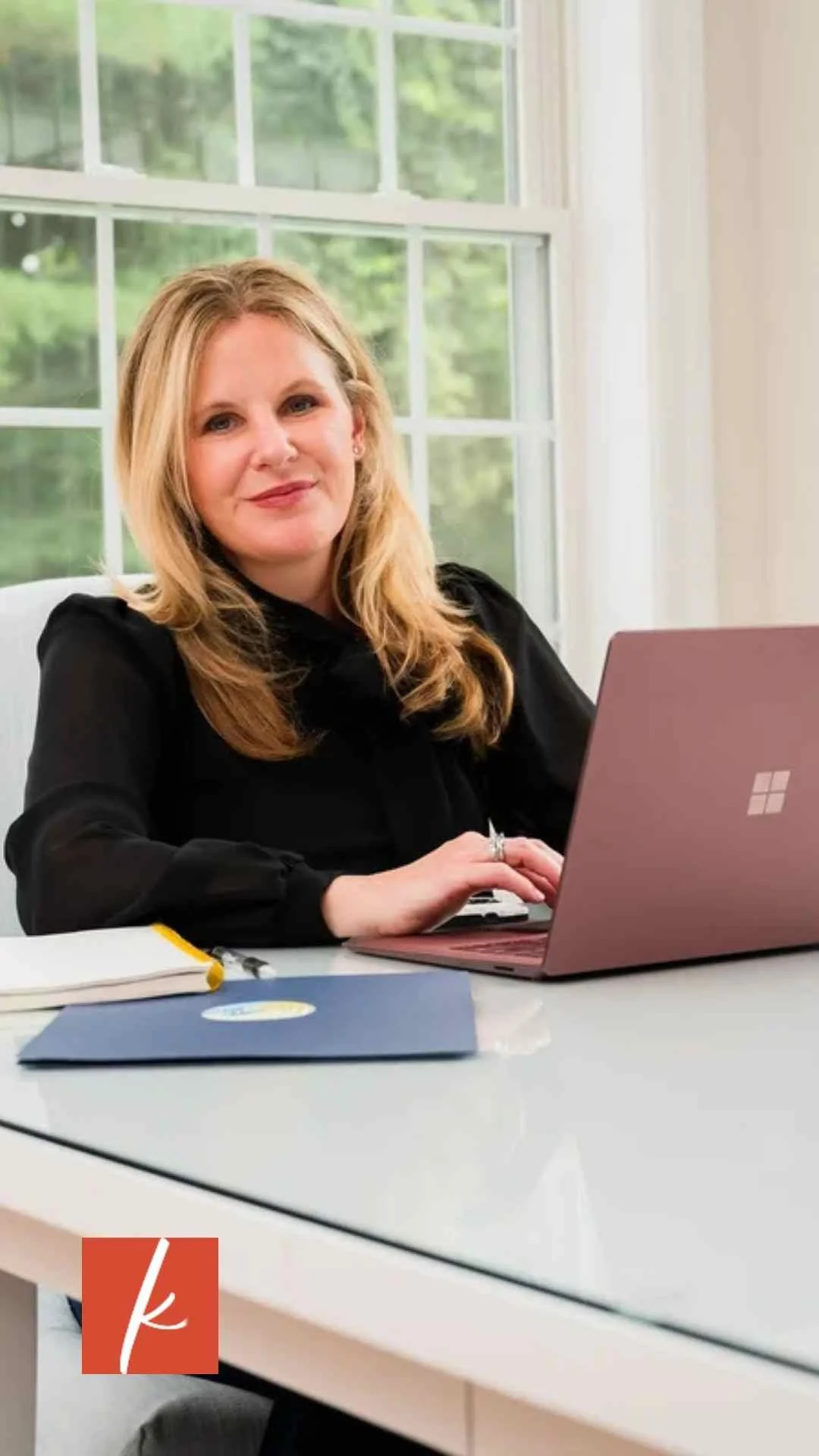 Woman seated at a desk working on a laptop, smiling toward the camera in a bright home office with large windows.