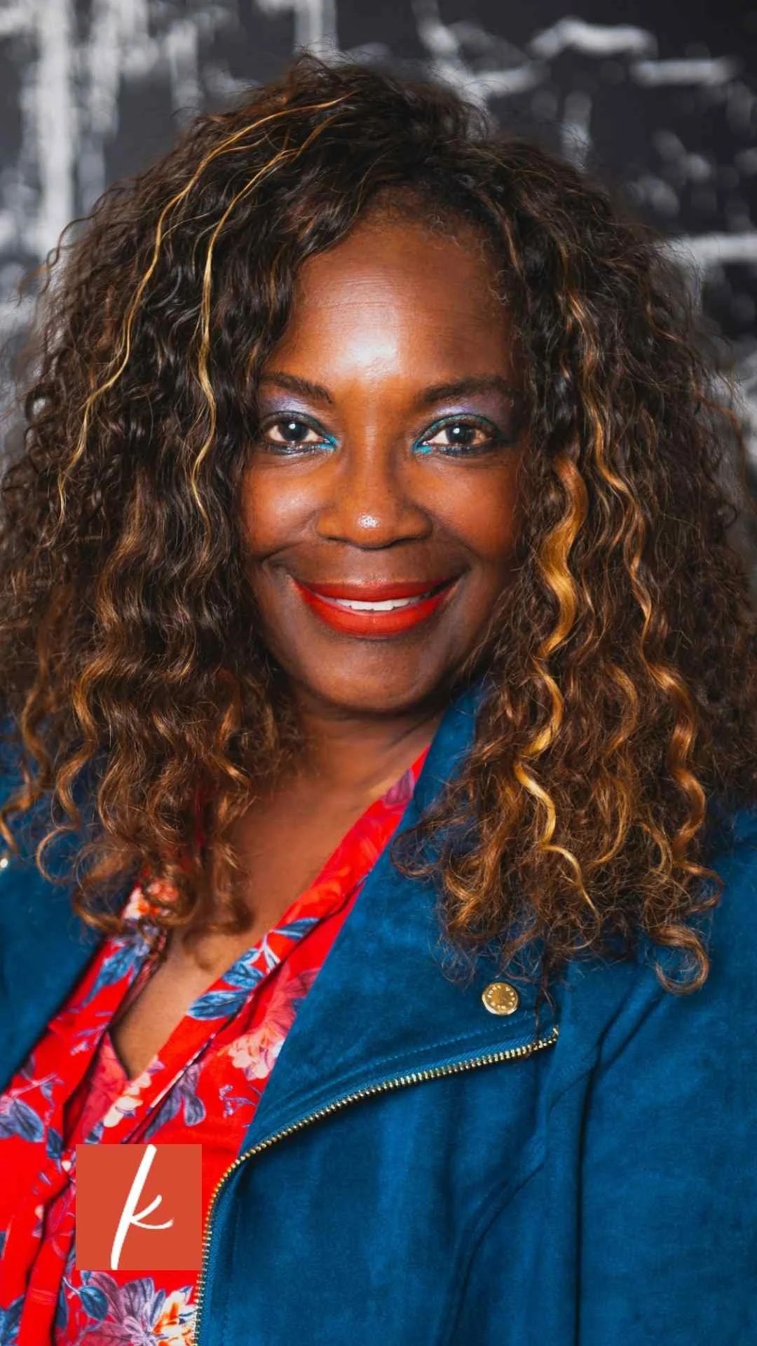 Smiling woman with curly highlighted hair wearing a teal jacket and red floral blouse, photographed against a dark textured background.