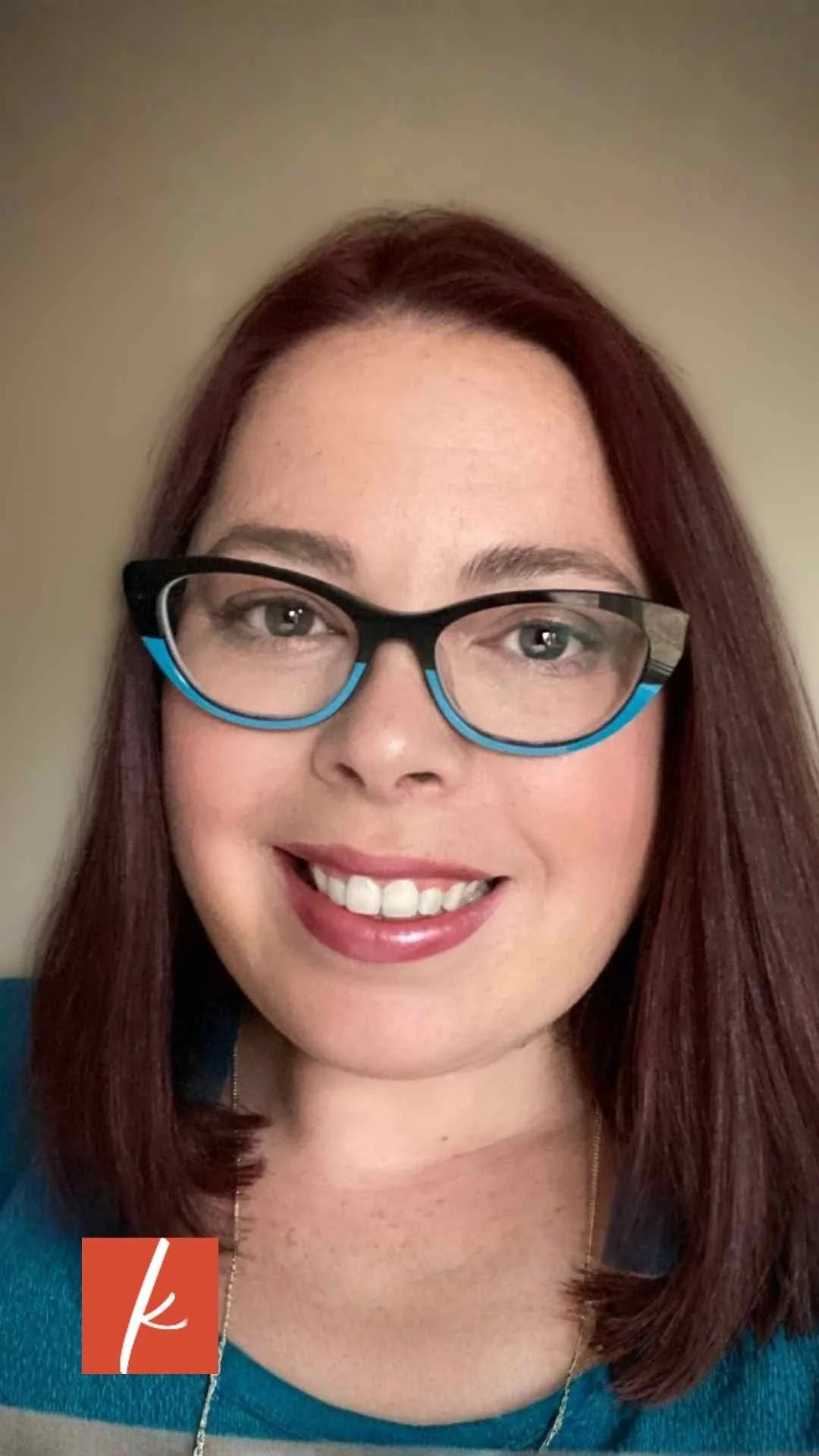 Close-up headshot of a smiling woman with red hair wearing blue-accented glasses and a teal top.