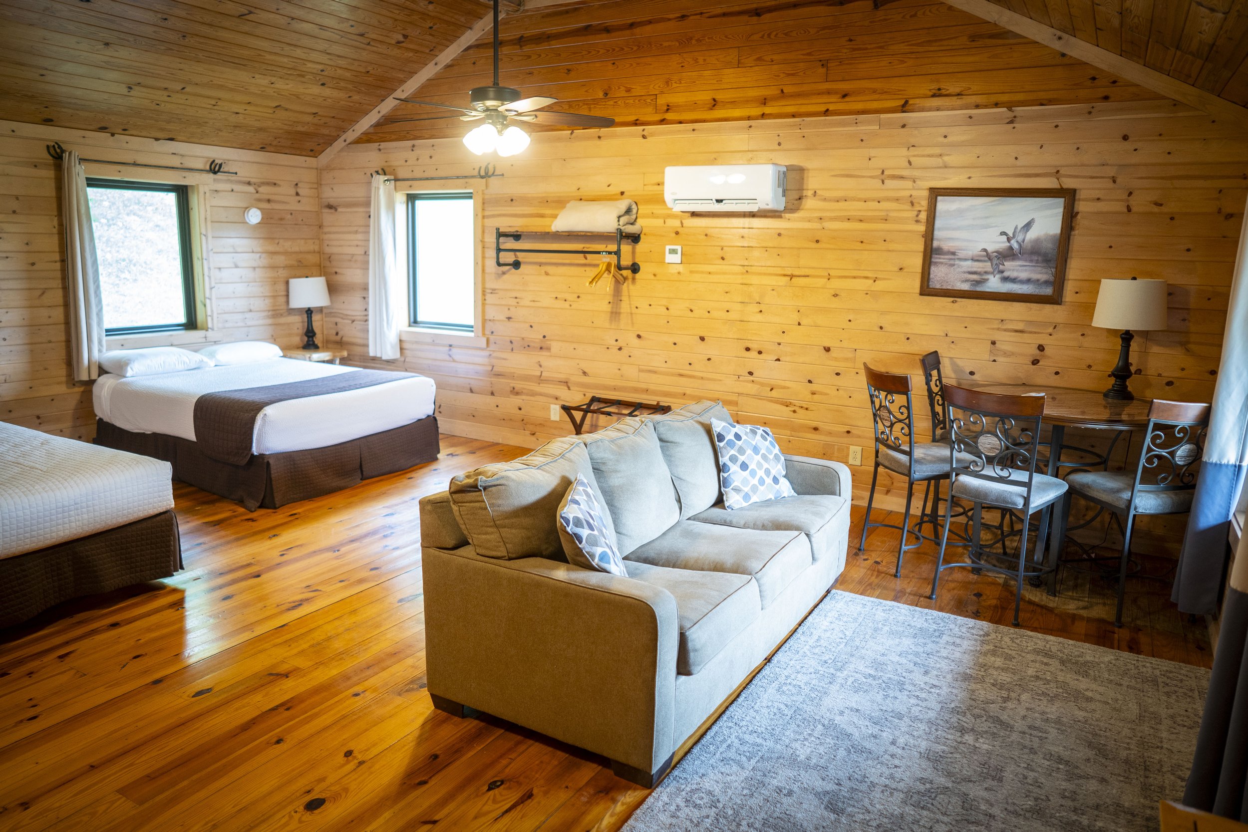 A cozy wooden interior with a bed, a sofa, a dining table with chairs, and windows, in a cabin-style room.