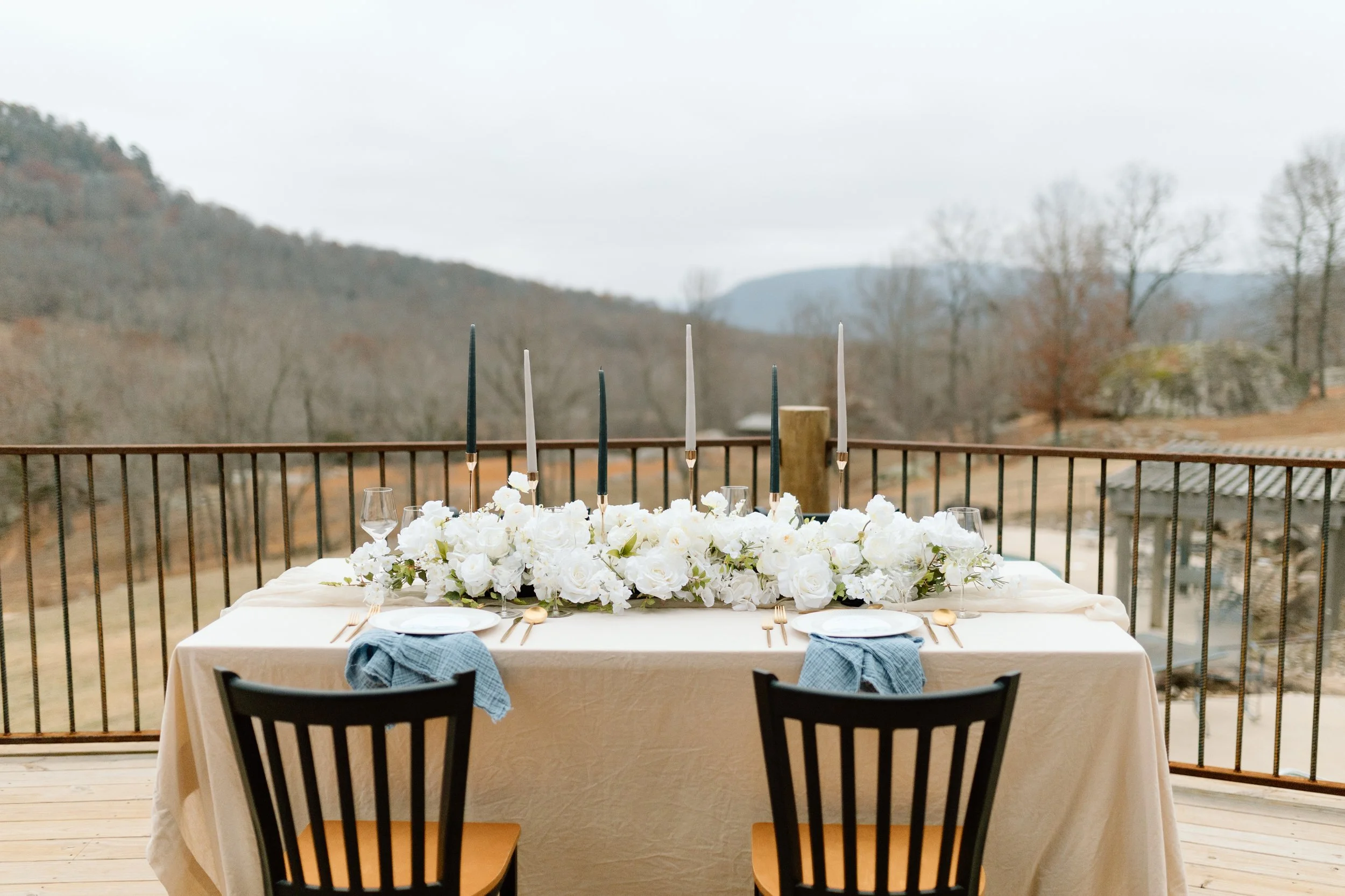 A table set outdoors with a white tablecloth, floral centerpiece of white flowers, black and white taper candles, white plates, gold flatware, and blue cloth napkins on black chairs, overlooking a rural landscape with trees and mountains.