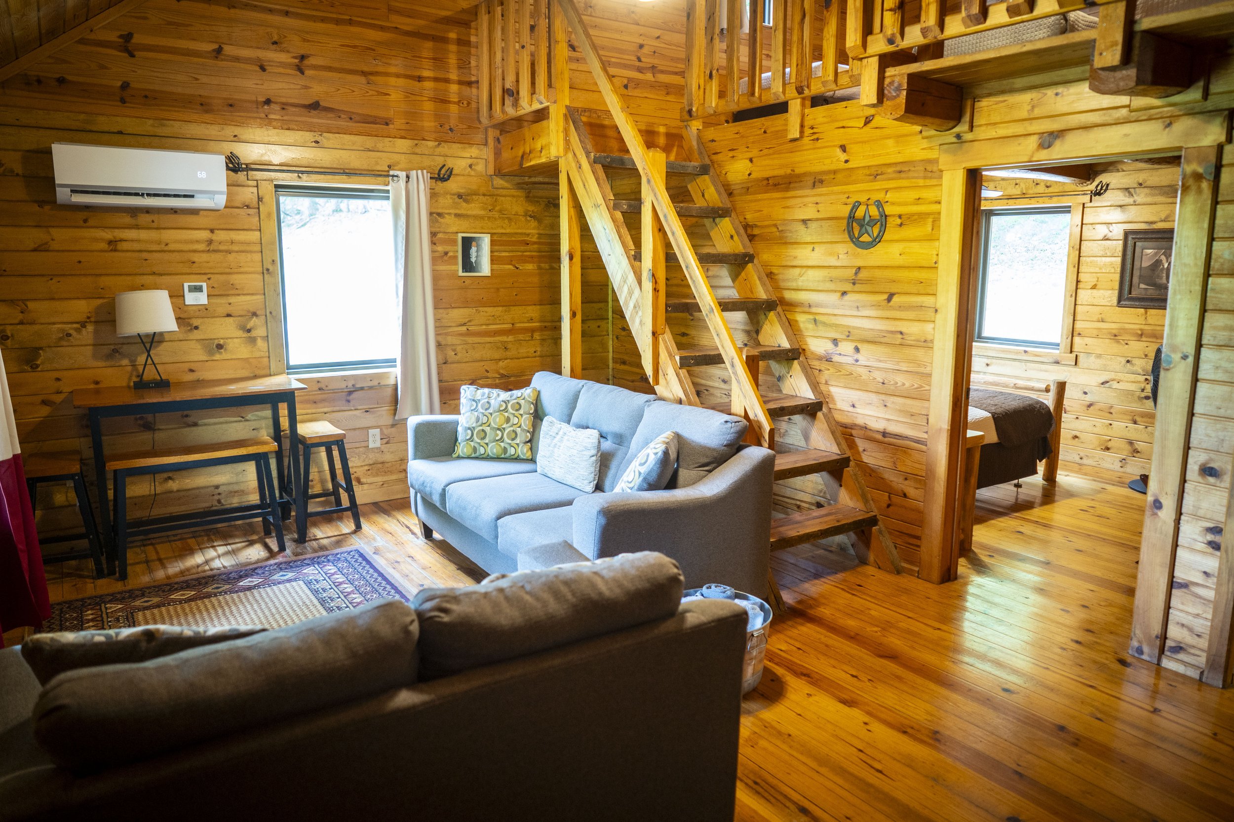 Interior of a cozy wooden cabin living room with a blue sofa, a wooden staircase, and a bedroom visible through an open doorway.