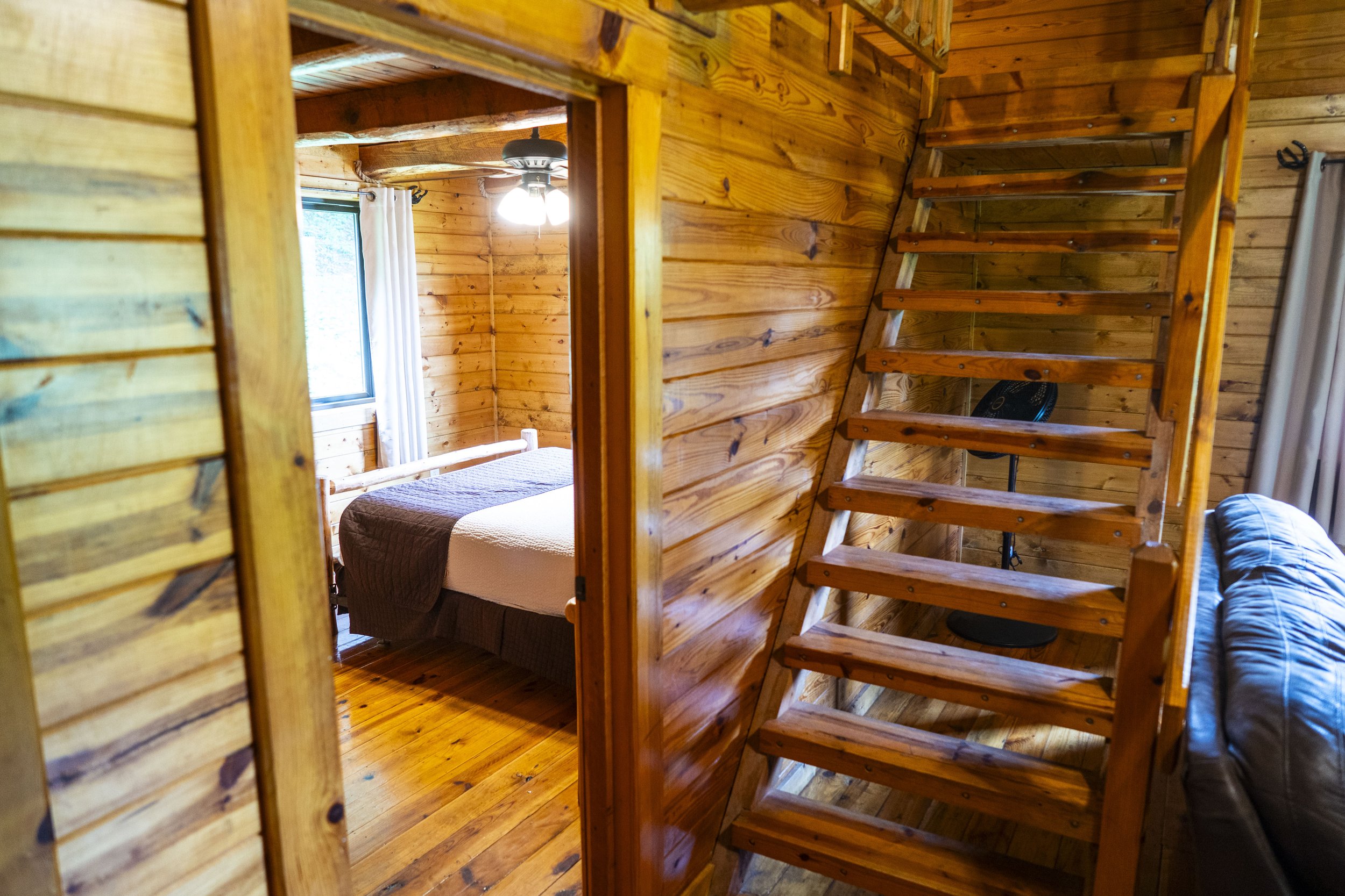 Interior view of a cozy wooden cabin with a bedroom visible through a doorway, featuring a bed with white and brown bedding, a window with white curtains, and a wooden staircase leading to an upper level.