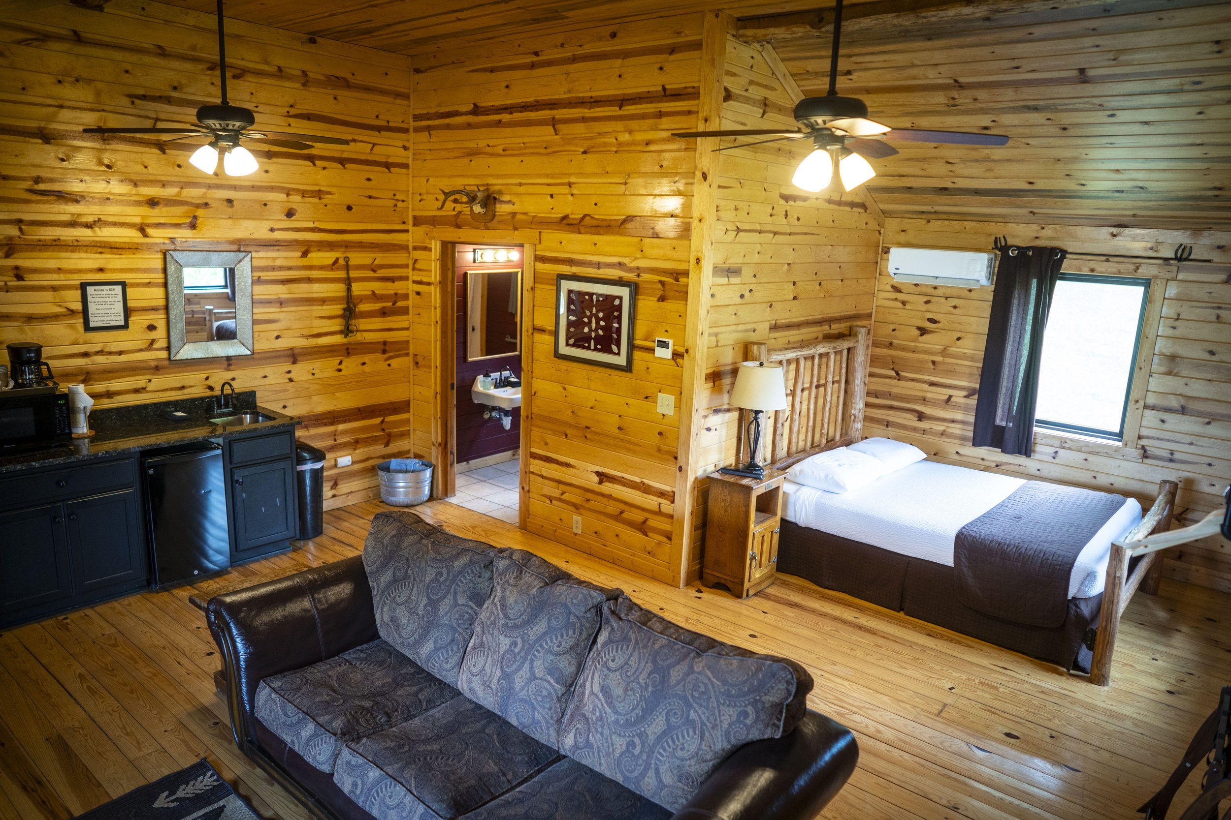 Interior of a cozy wooden cabin with a bed, a sofa, a kitchen area, and a doorway to a bathroom, illuminated by ceiling fans and natural light from windows.