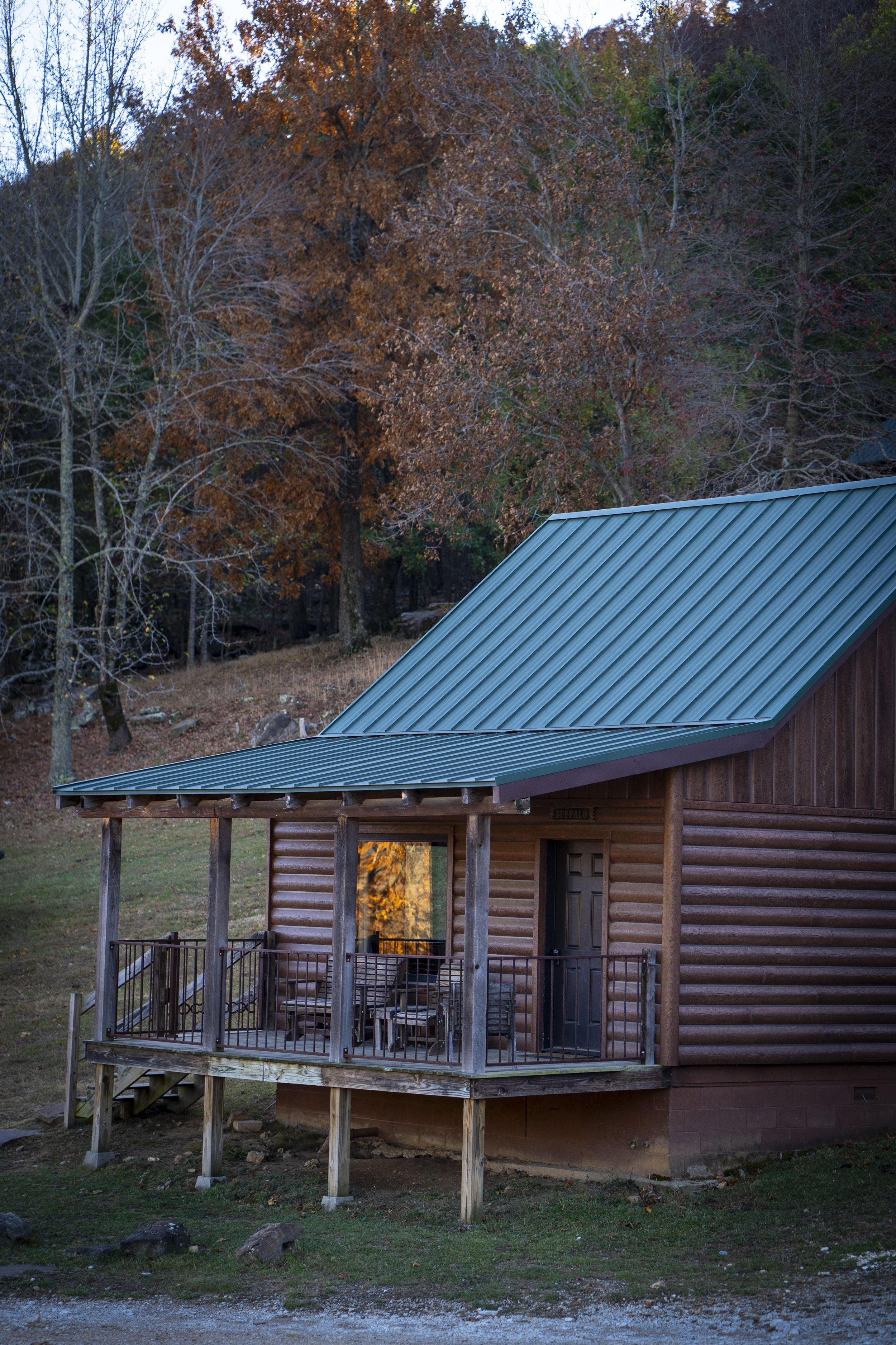 A log cabin with a green metal roof and a small porch with chairs, situated in a wooded area with trees showing autumn colors.