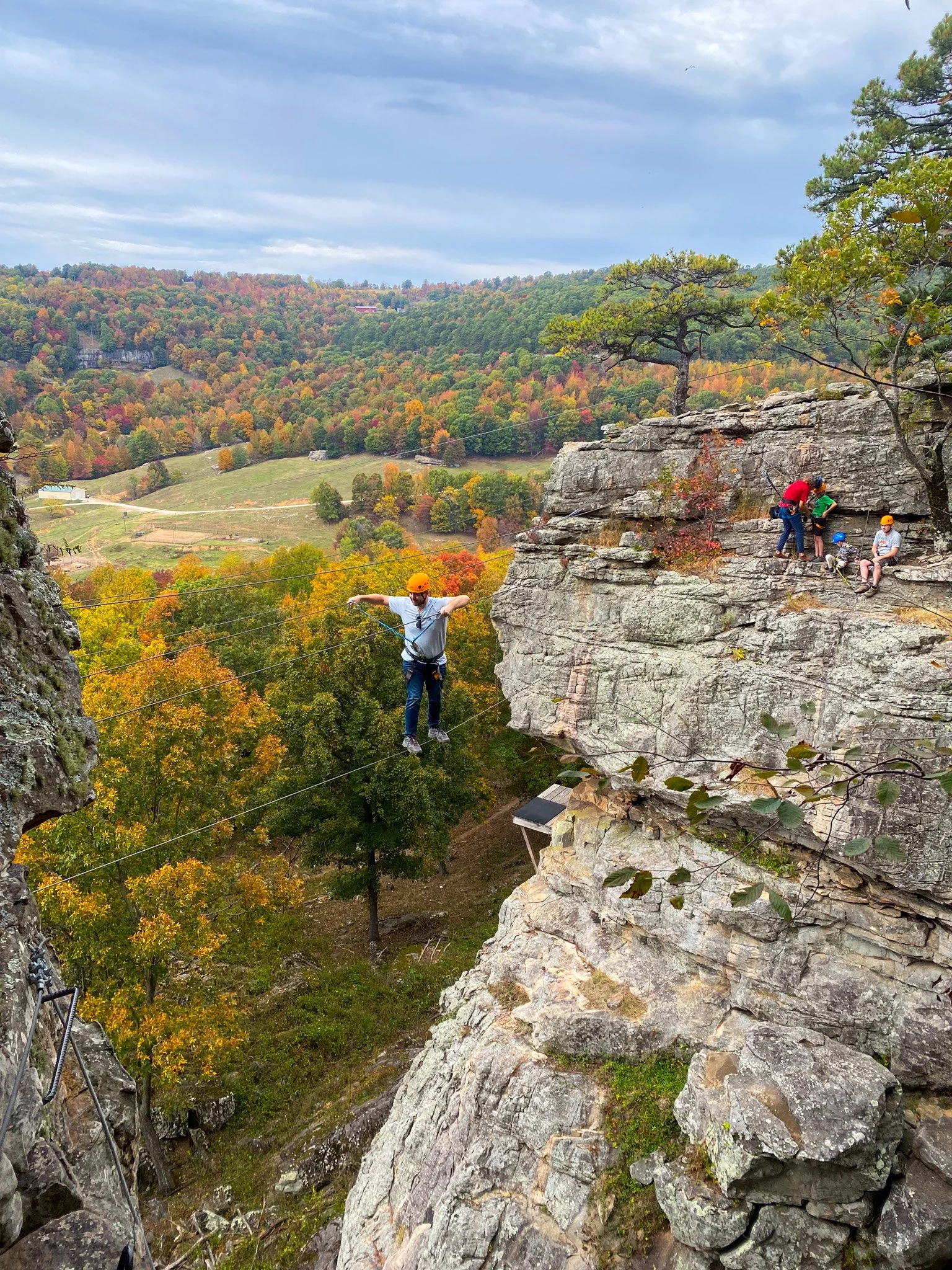 A person walking on a tightrope high above the ground with a safety harness, with a group of people nearby on the rocky cliff edge, surrounded by a scenic valley with trees displaying fall foliage.