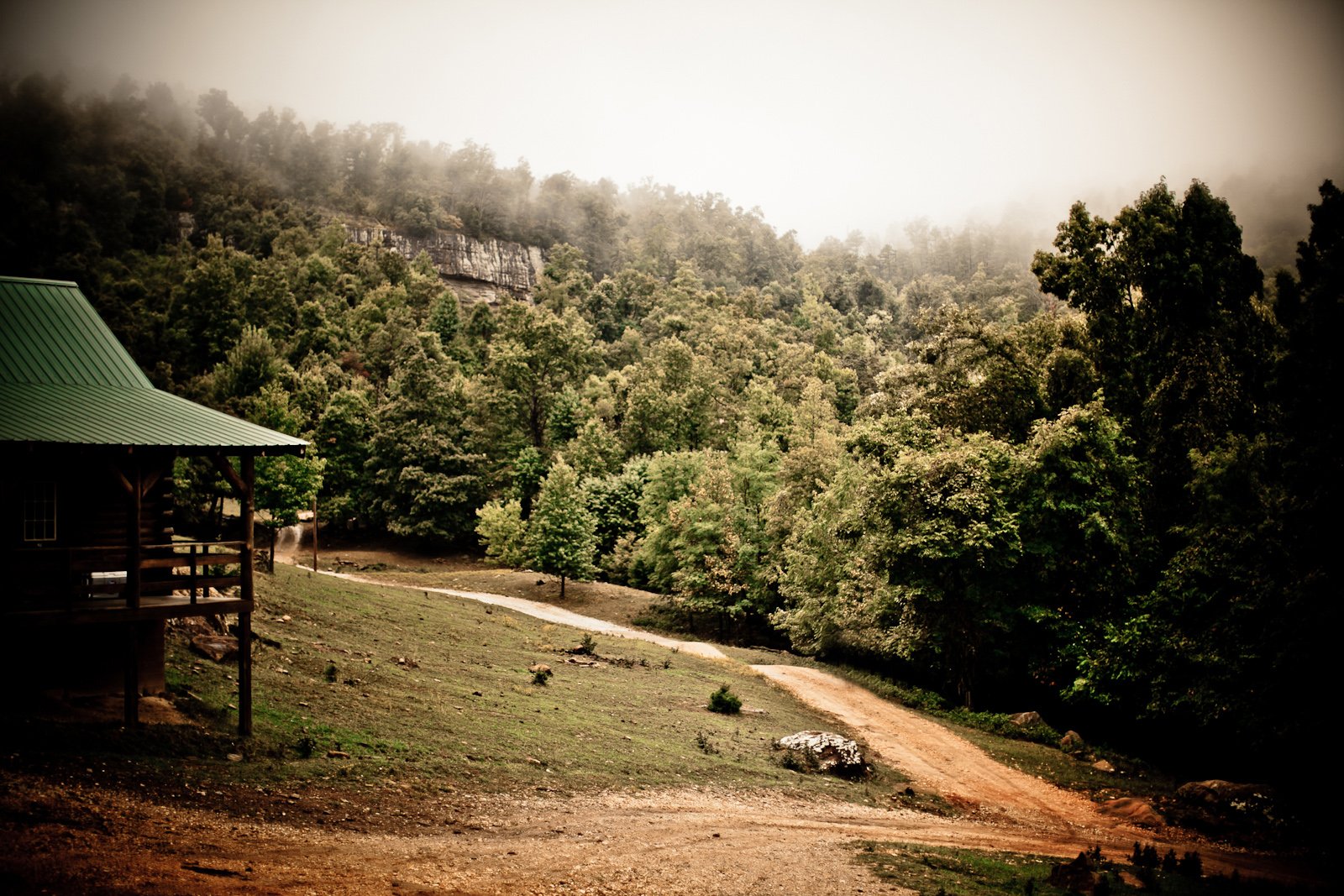 A dirt path running through a wooded hillside with a house with a green roof on the left, foggy mountains in the background.