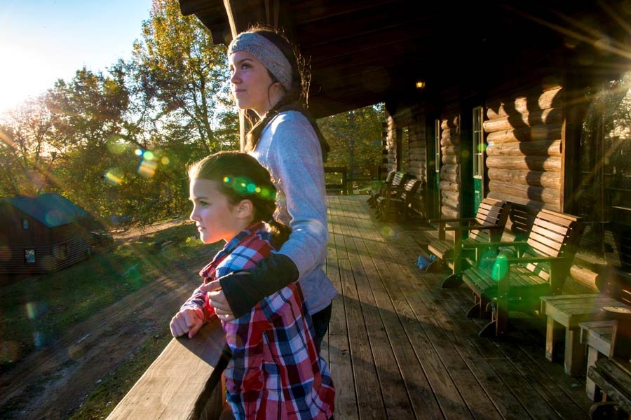 Two young girls stand on a wooden porch of a log cabin at sunset, looking into the distance. One girl is older, wearing a headband and a white sweater, while the younger girl is wearing a red and blue plaid shirt. Sunlight creates lens flares and ill