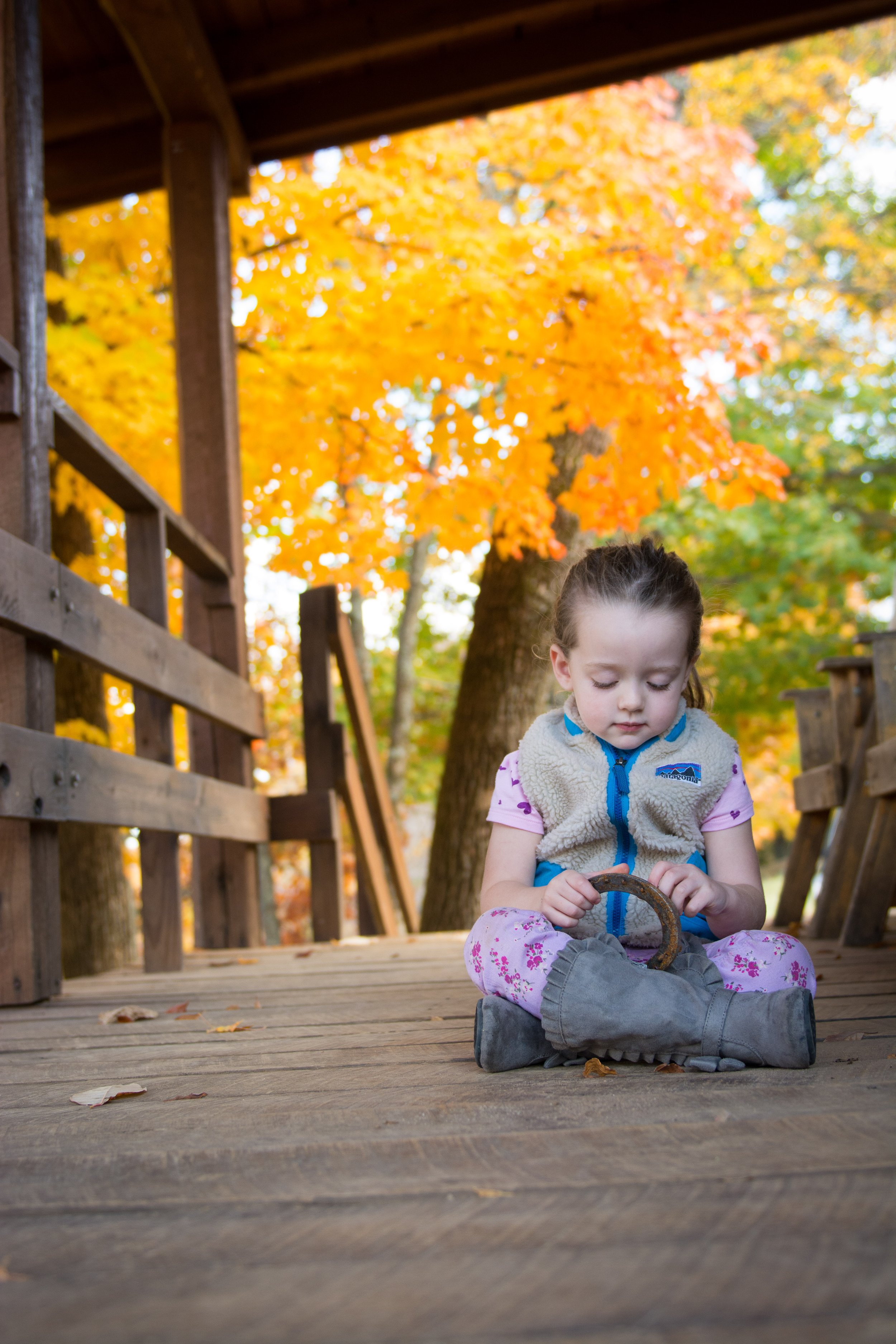 A young girl sitting on a wooden bridge in autumn, holding a horseshoe while looking down, with colorful fall leaves and trees in the background.