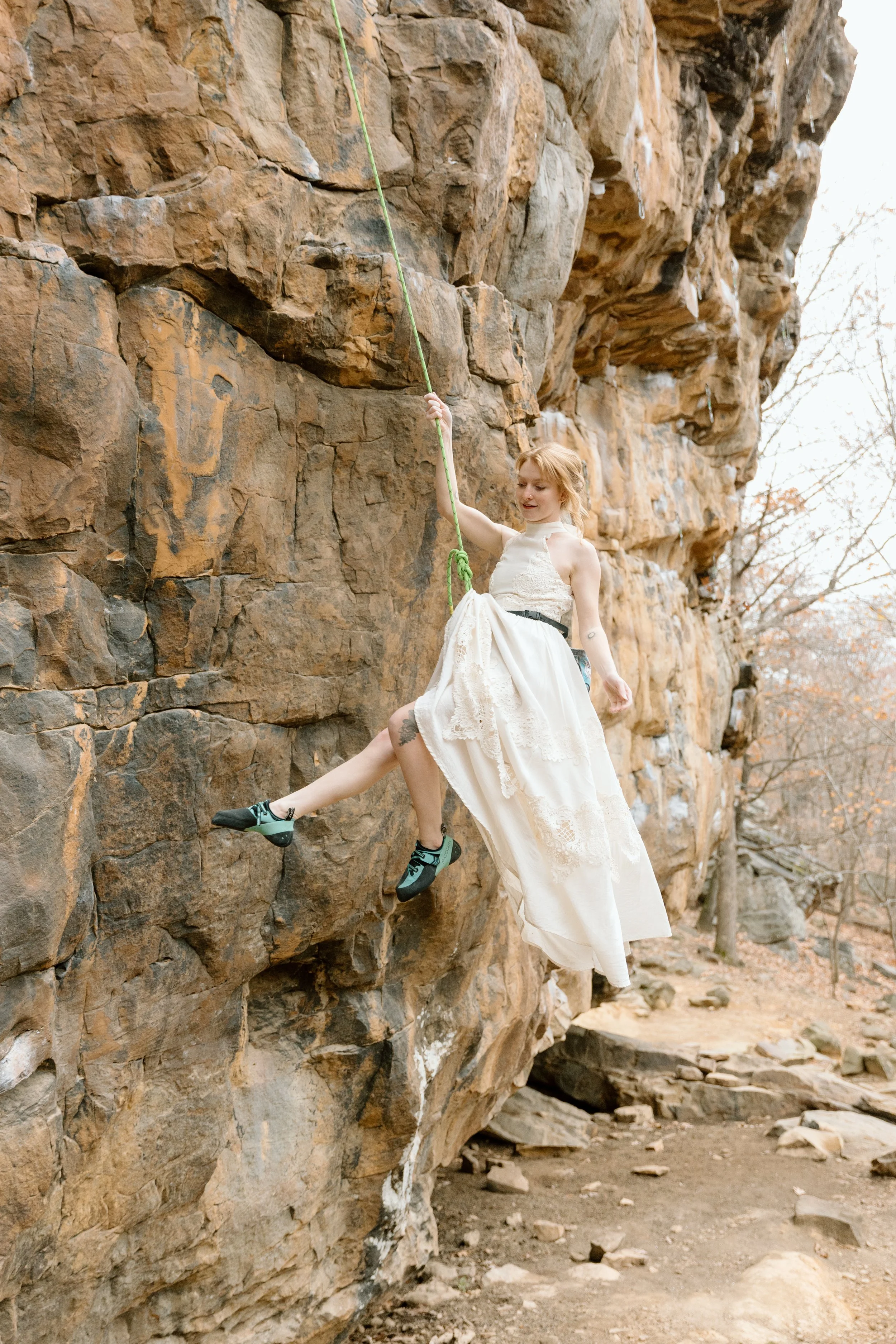 A woman in a white lace dress is rock climbing outdoors on a large brown rock formation, using a green climbing rope for support.