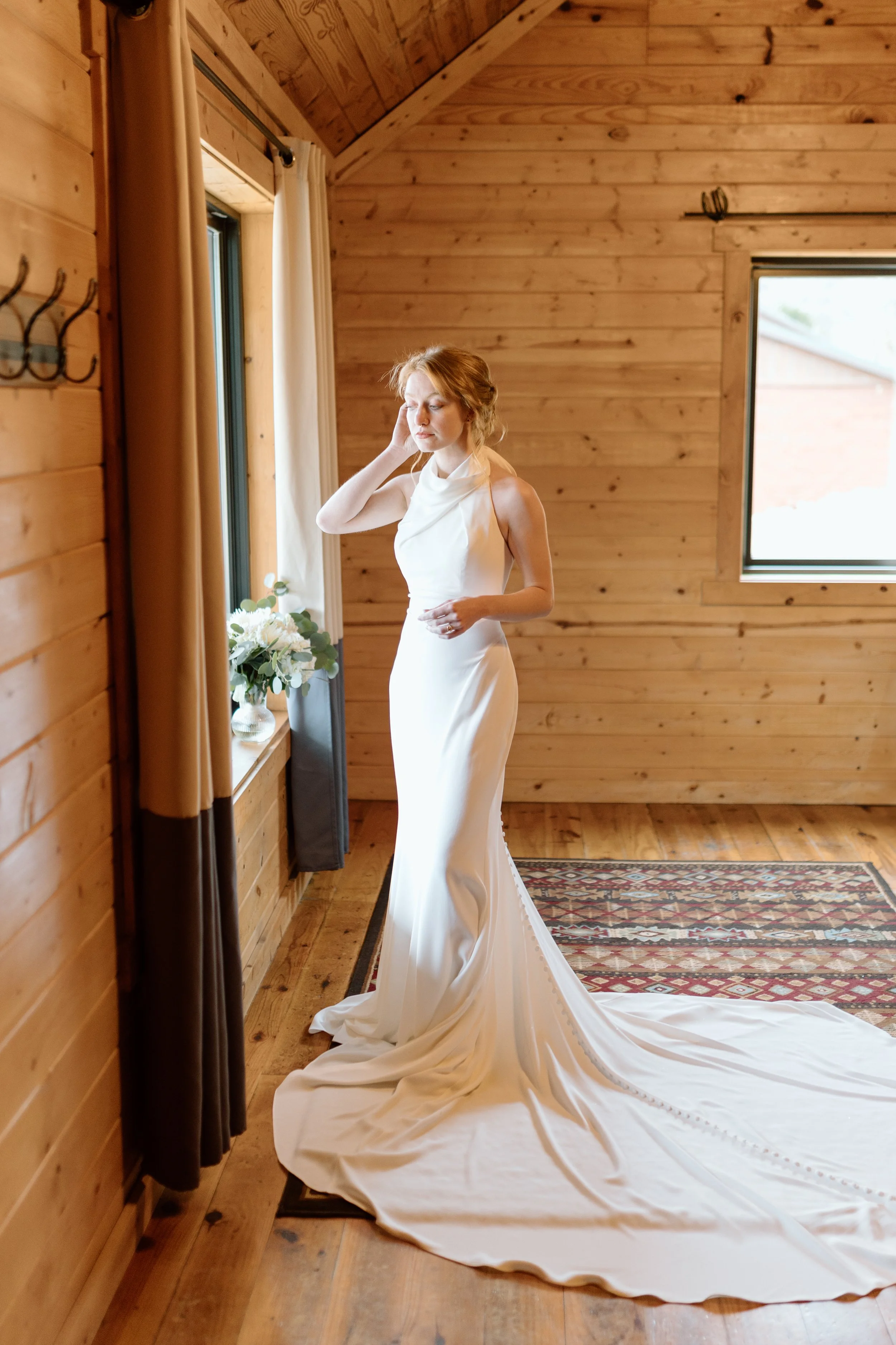 A woman in a white wedding gown standing in a wooden room by a window, touching her face and looking contemplative.