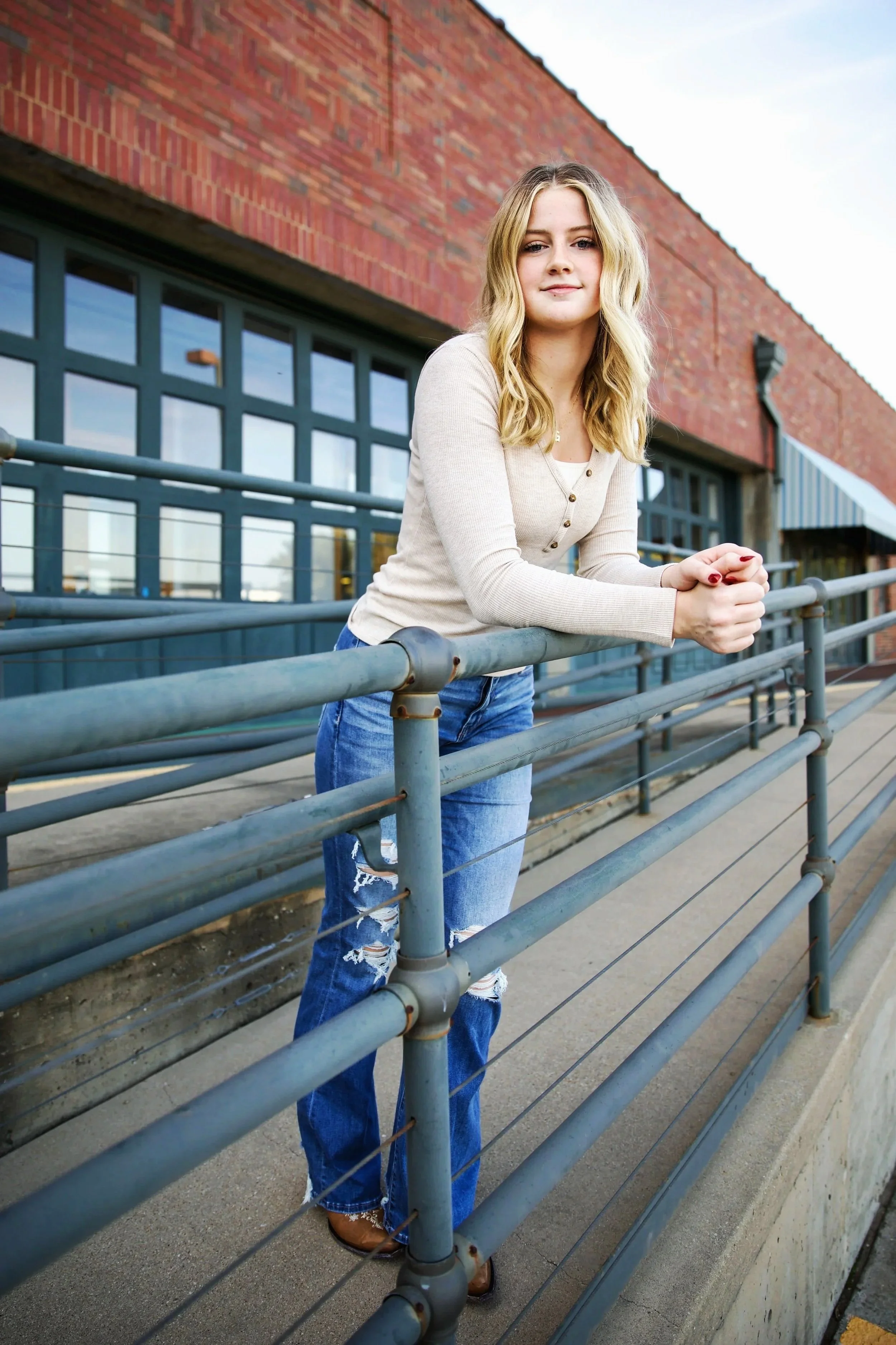 A young woman with blonde hair and a neutral expression, wearing a beige long-sleeved shirt and ripped jeans, leaning on a metal railing outside a brick building with large windows. Individual senior photo session at UTA Fort Worth. 