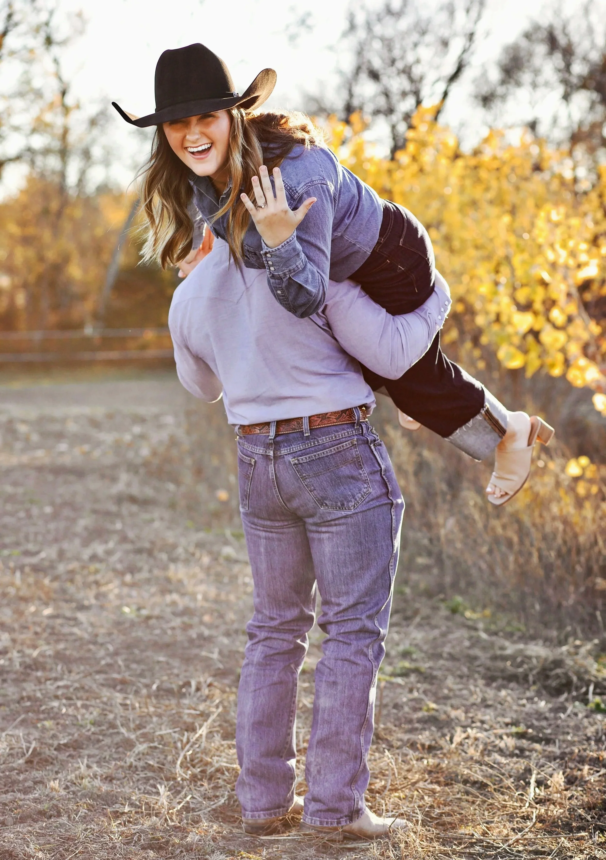 A woman with long hair wearing a cowboy hat, denim jacket, and jeans, is smiling and being piggybacked by her fiancee while showing off her engagement ring outdoors on a sunny day during fall with trees showing yellow leaves.