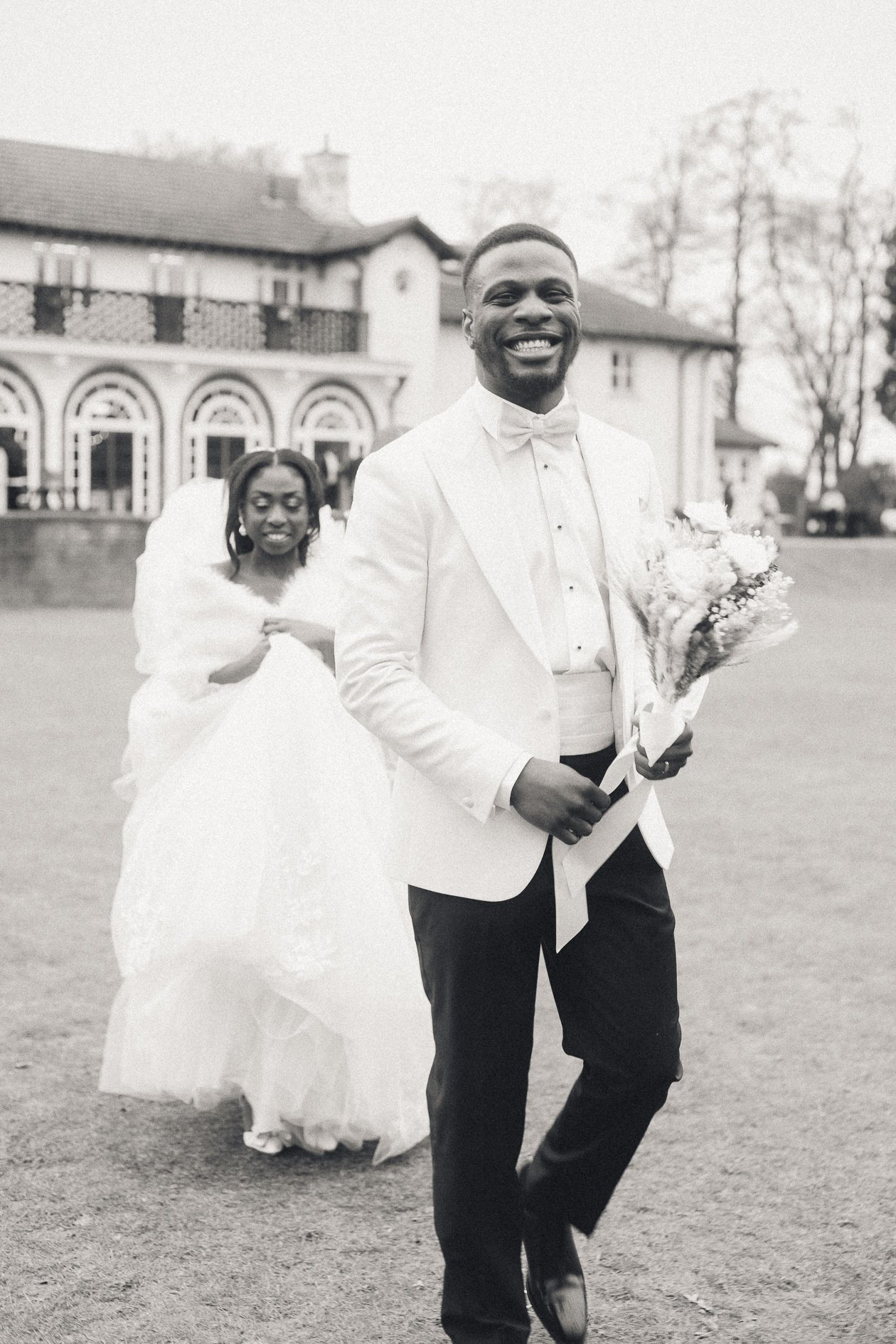 Black man in a white suit and bow tie holding a bouquet of flowers, smiling, with a woman in a wedding dress behind him, outside in front of a house.
