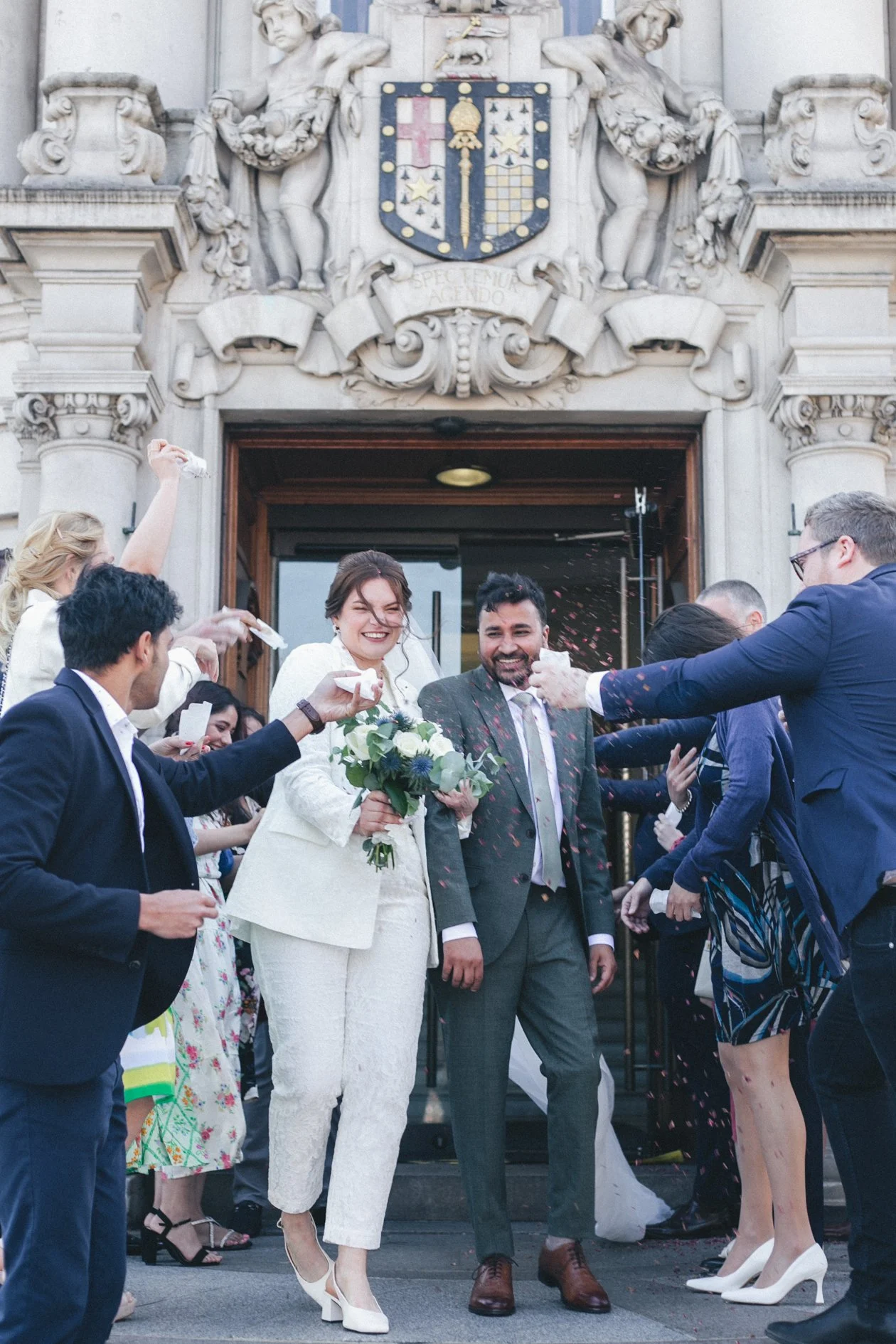 A newly married couple exiting Lambeth Town Hall, surrounded by guests throwing confetti and cheering. The bride holds a bouquet of flowers, and both are smiling. The building has an ornate stone facade with a crest above the entrance.