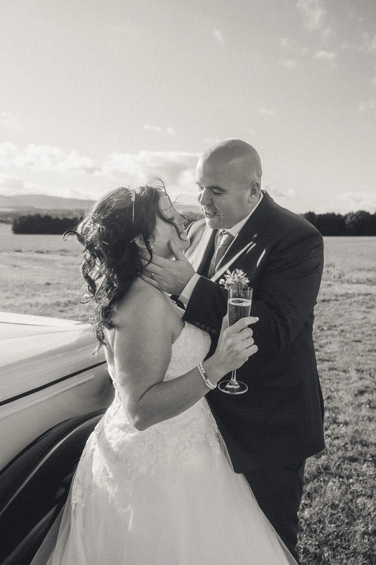 Black and white photo of a bride and groom outdoors, with the groom gently holding the bride's face. The bride is holding a champagne glass, and they are standing near a vintage car with a scenic landscape in the background.
