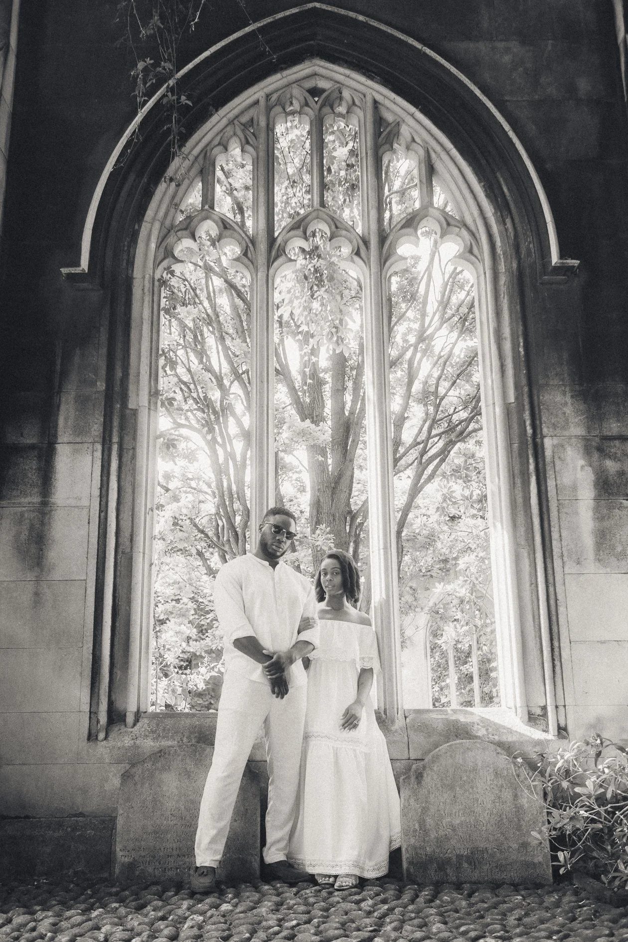 A man and woman in white clothing standing in front of a large Gothic-style arched window with trees visible outside.