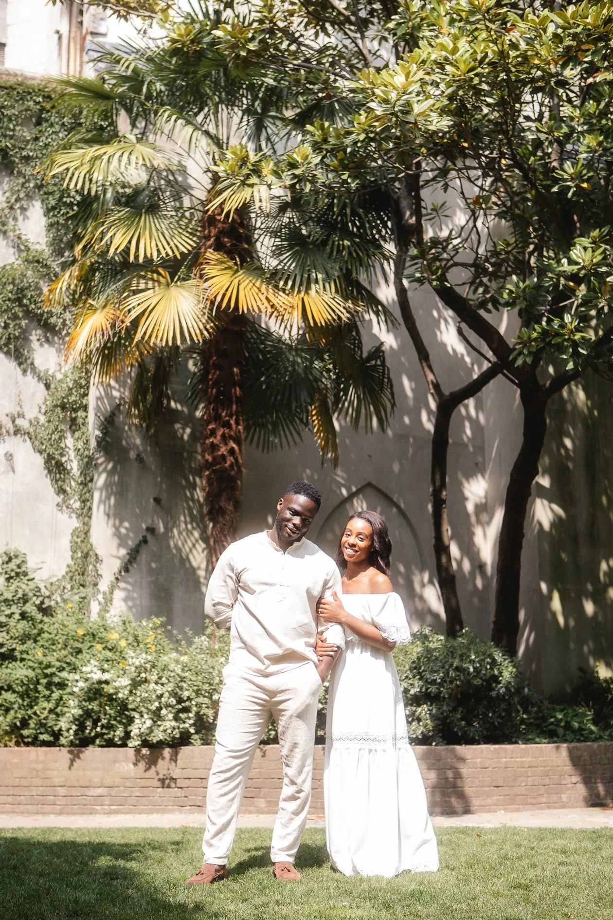 A smiling couple stands on a lawn in a garden with lush palm and other trees, and a white wall in the background.