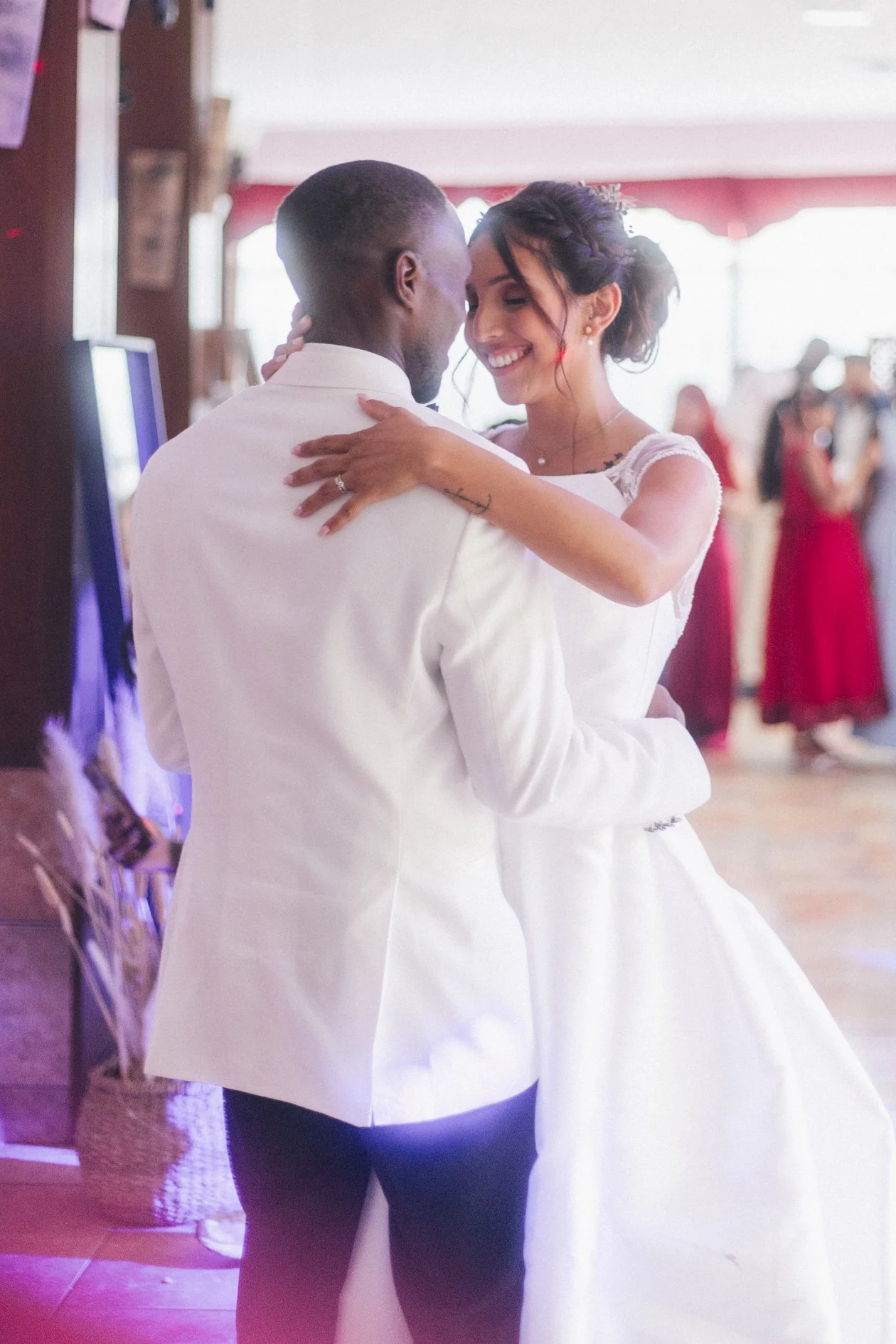 A newlywed couple dancing together, smiling, at their wedding reception. The bride is dressed in a white wedding gown, and the groom is in a white suit jacket.