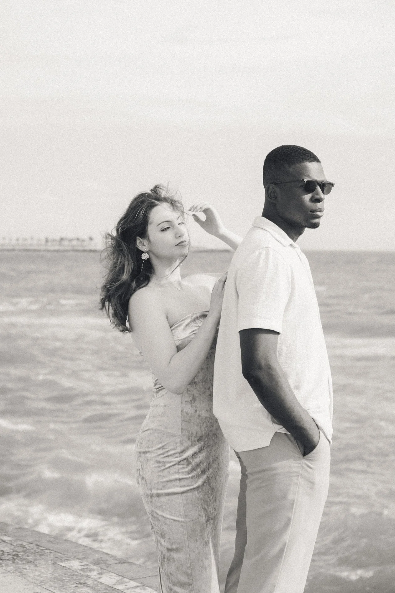 A black and white photo of a man and woman standing by the ocean. The woman is wearing a strapless dress and earrings, and the man is wearing a short-sleeved shirt and sunglasses. The woman is touching the man's shoulder while the man looks away.