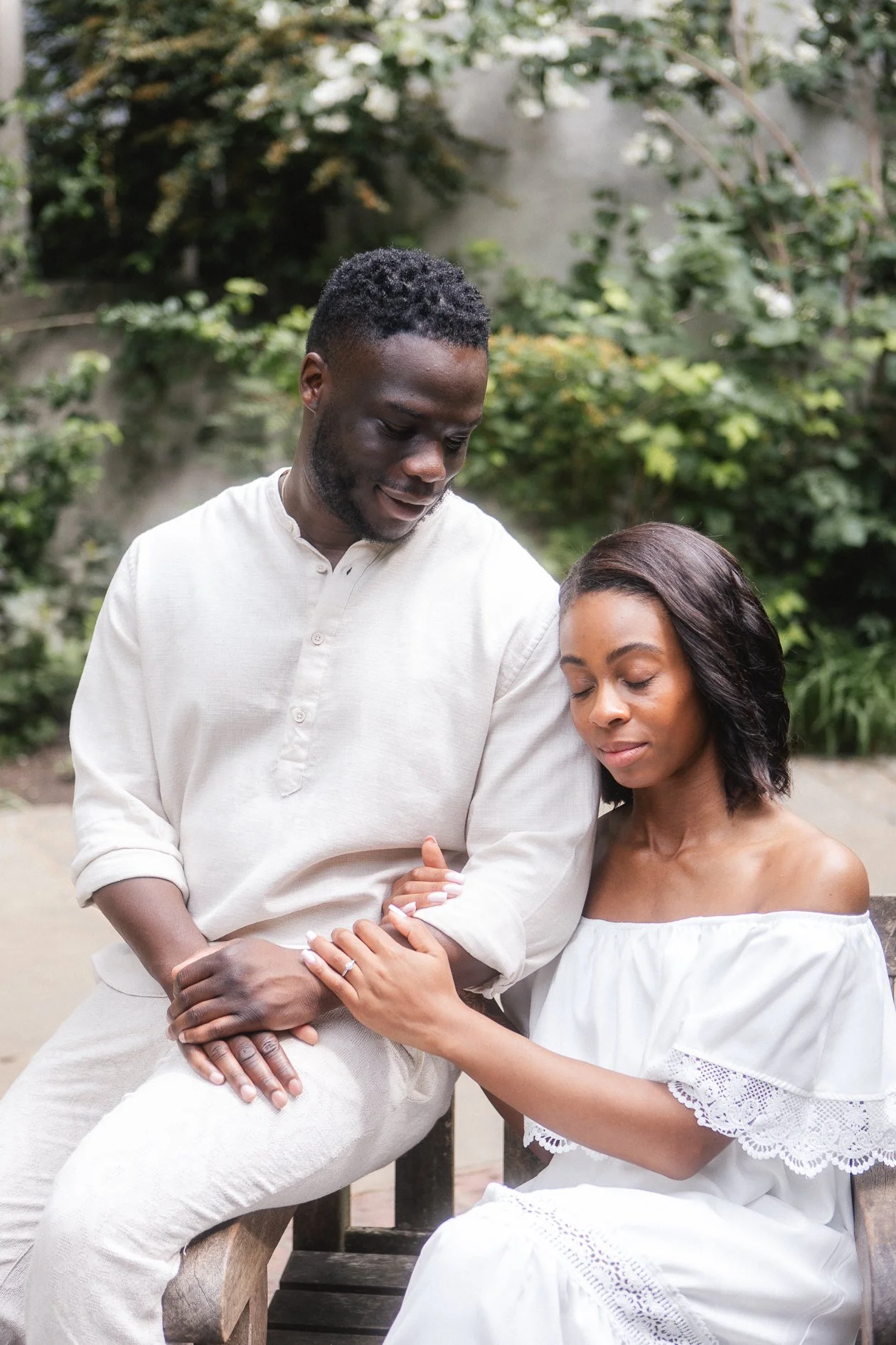 A couple sitting outdoors, with the man gently holding the woman's hand on her lap. The woman has her eyes closed and appears peaceful, while the man is looking down at her. Both are wearing white clothing, and there is greenery in the background.