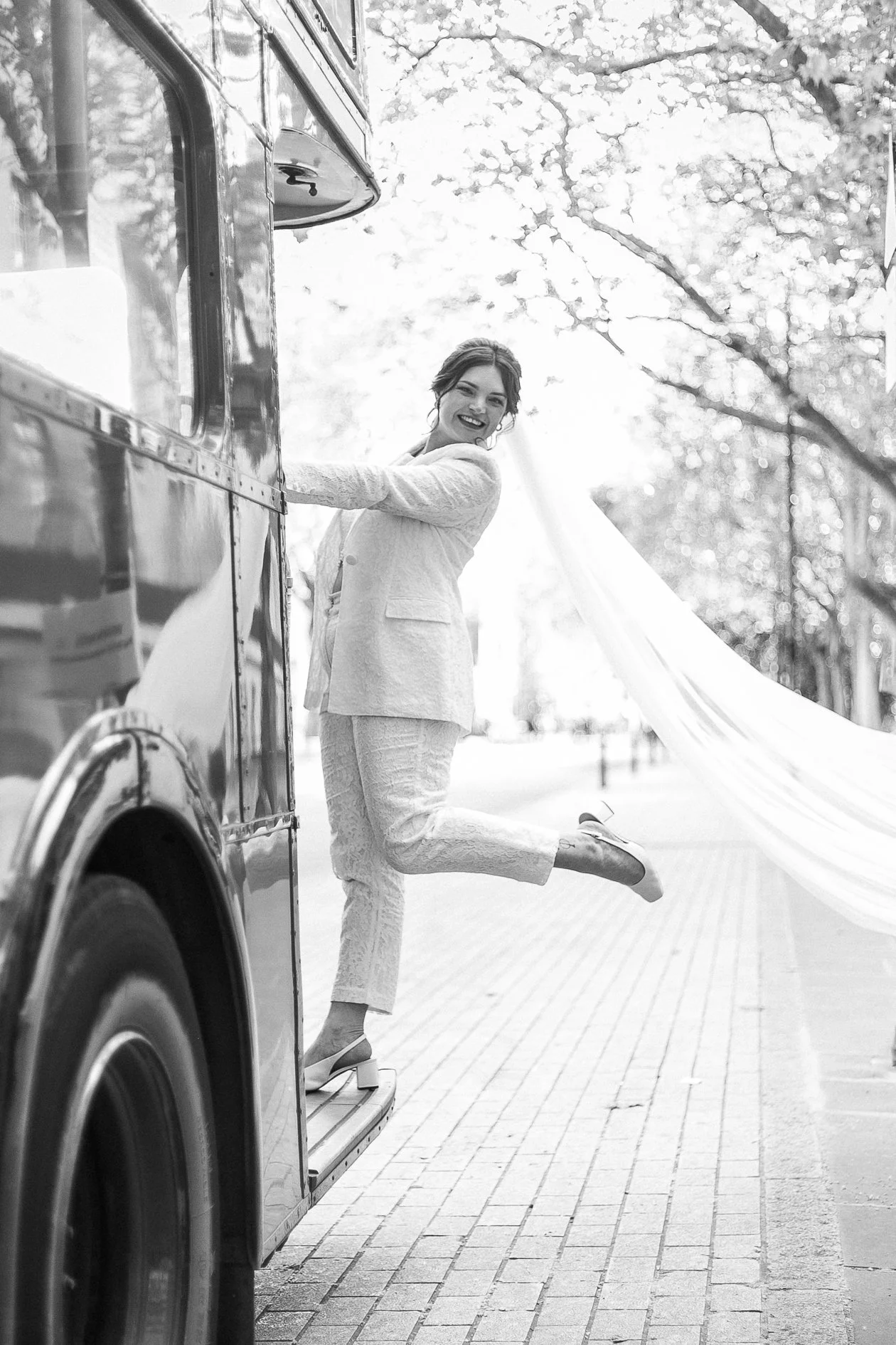 A woman in a light-colored suit standing on the steps of a vintage London bus, smiling and playfully kicking one leg in a park with trees and a fabric canopy in the background, captured in black and white.