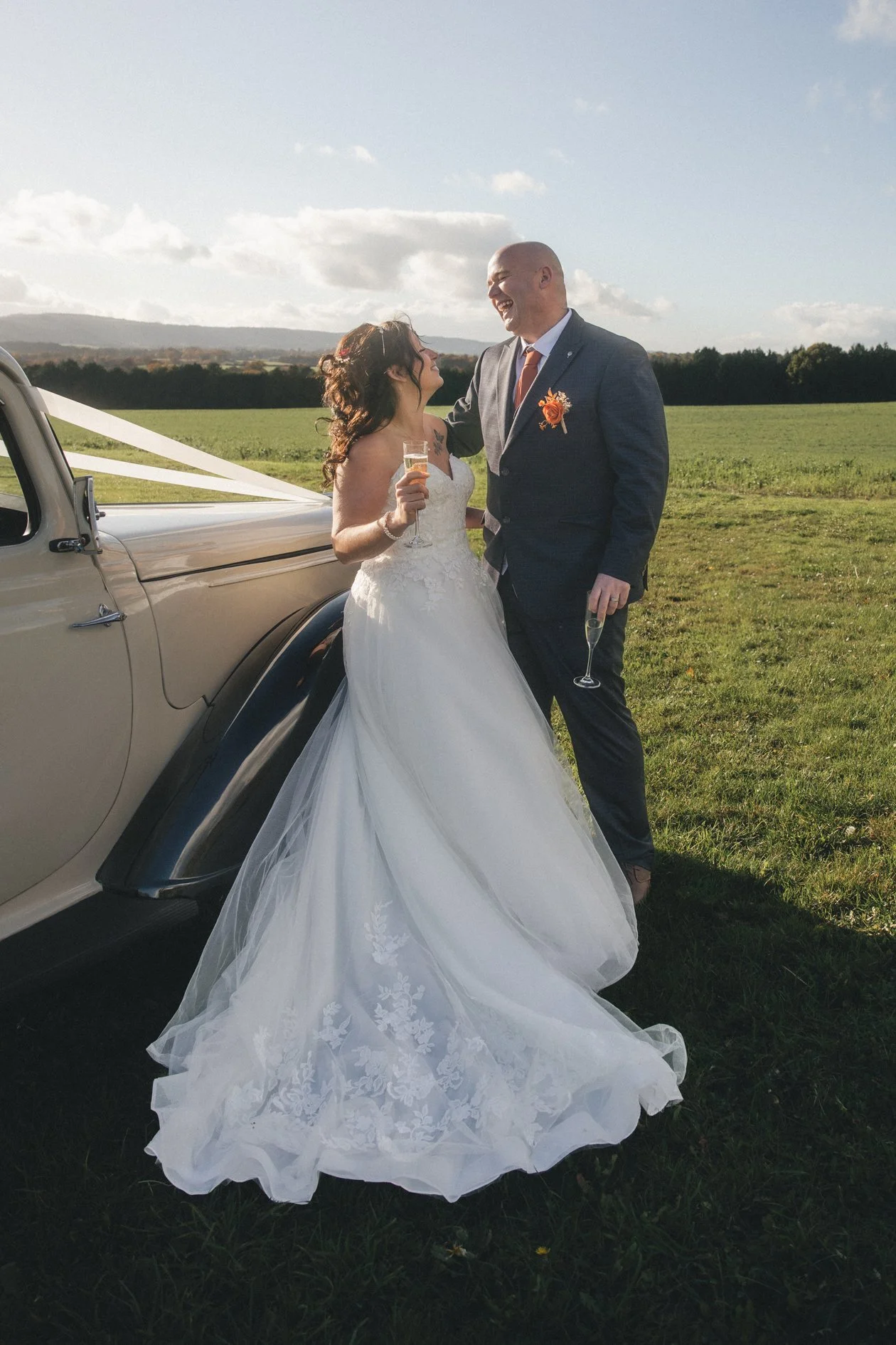 Bride and groom laughing and celebrating with champagne outdoors in a field, beside a vintage car decorated with white ribbons.