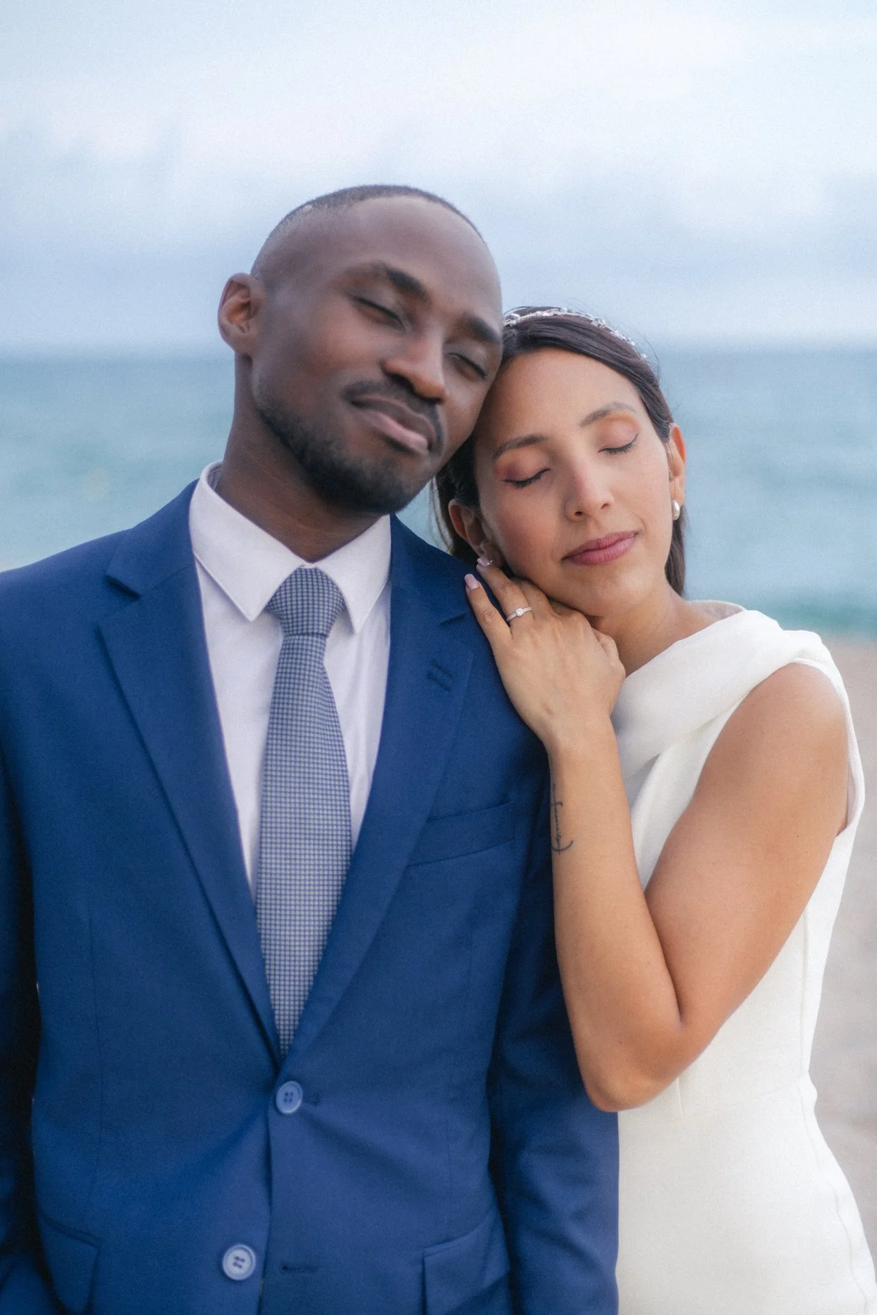 A smiling man in a blue suit and a woman in a white dress with closed eyes, standing close together with the woman resting her head on the man's shoulder at the beach.