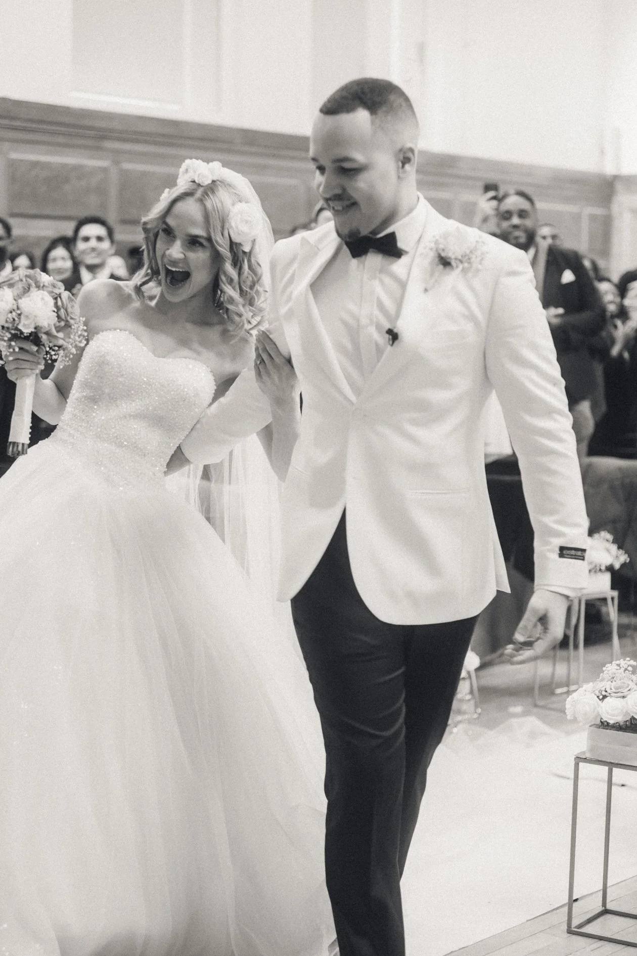 A bride and groom exchanging rings during their wedding ceremony, surrounded by guests; the bride is wearing a wedding gown, and the groom is in a white tuxedo with a black bow tie, in a black and white photograph.