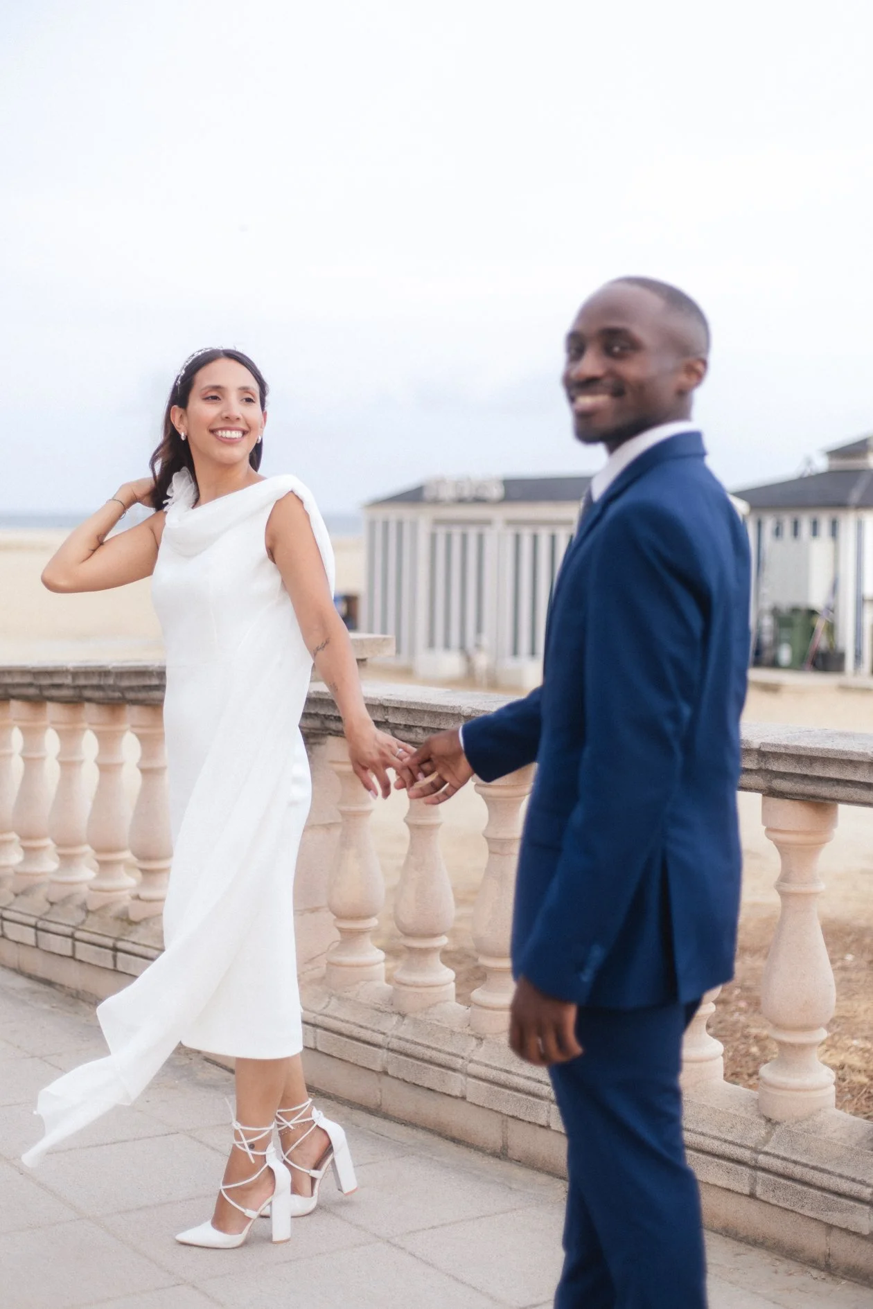 A couple holding hands and smiling at each other on a seaside promenade, with a beach and beach huts in the background, during daytime.