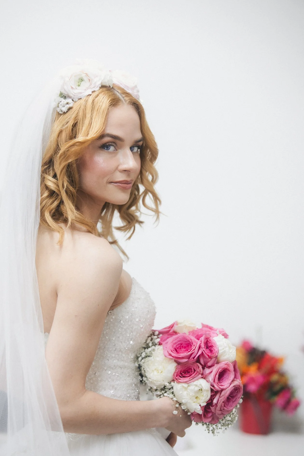 A bride with red hair, wearing a white wedding dress and veil, holding a bouquet of pink and white roses, standing against a plain white wall.