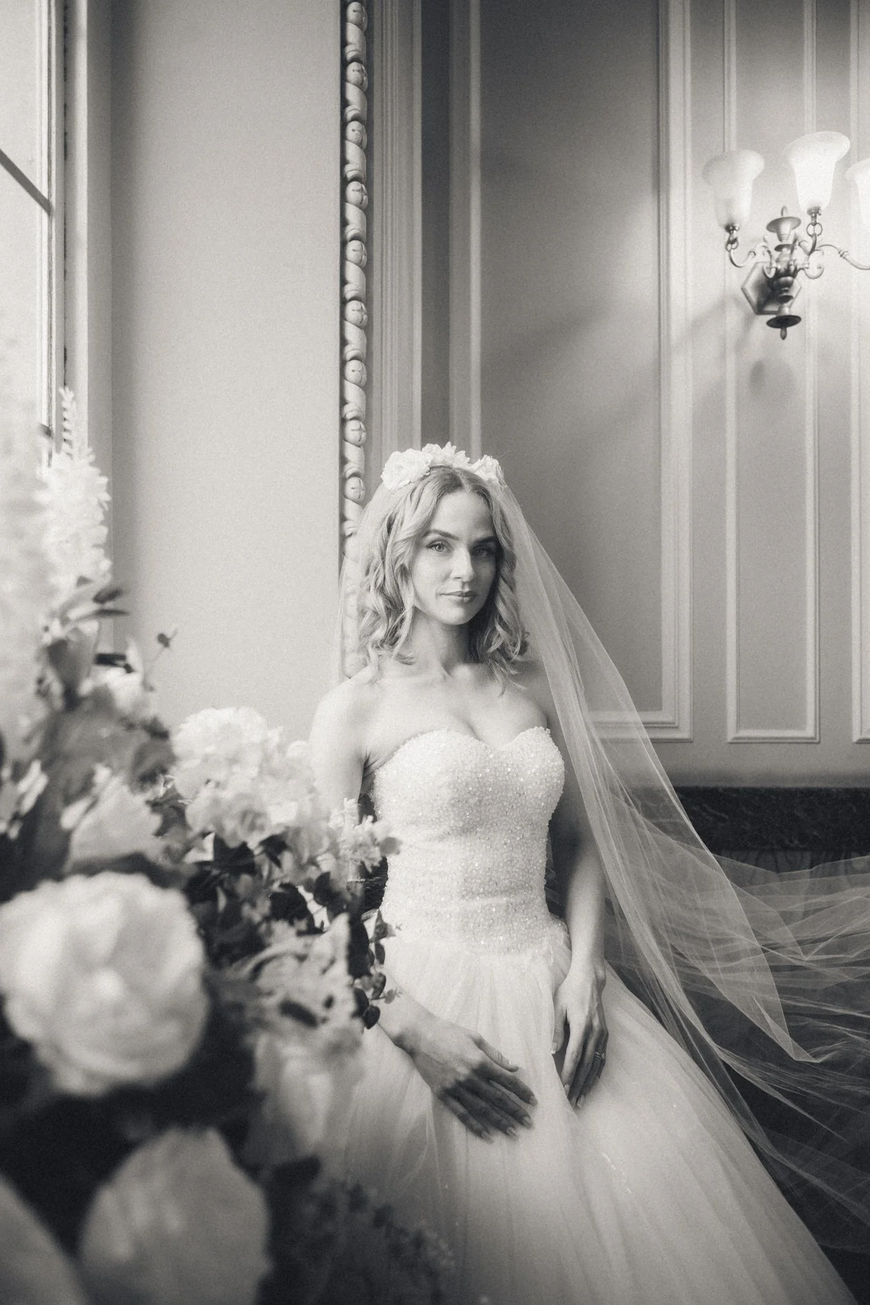 A bride in a strapless wedding gown with a tulle skirt, wearing a veil and a floral headpiece, sitting next to a bouquet of flowers in an elegant room with wall paneling and a chandelier.