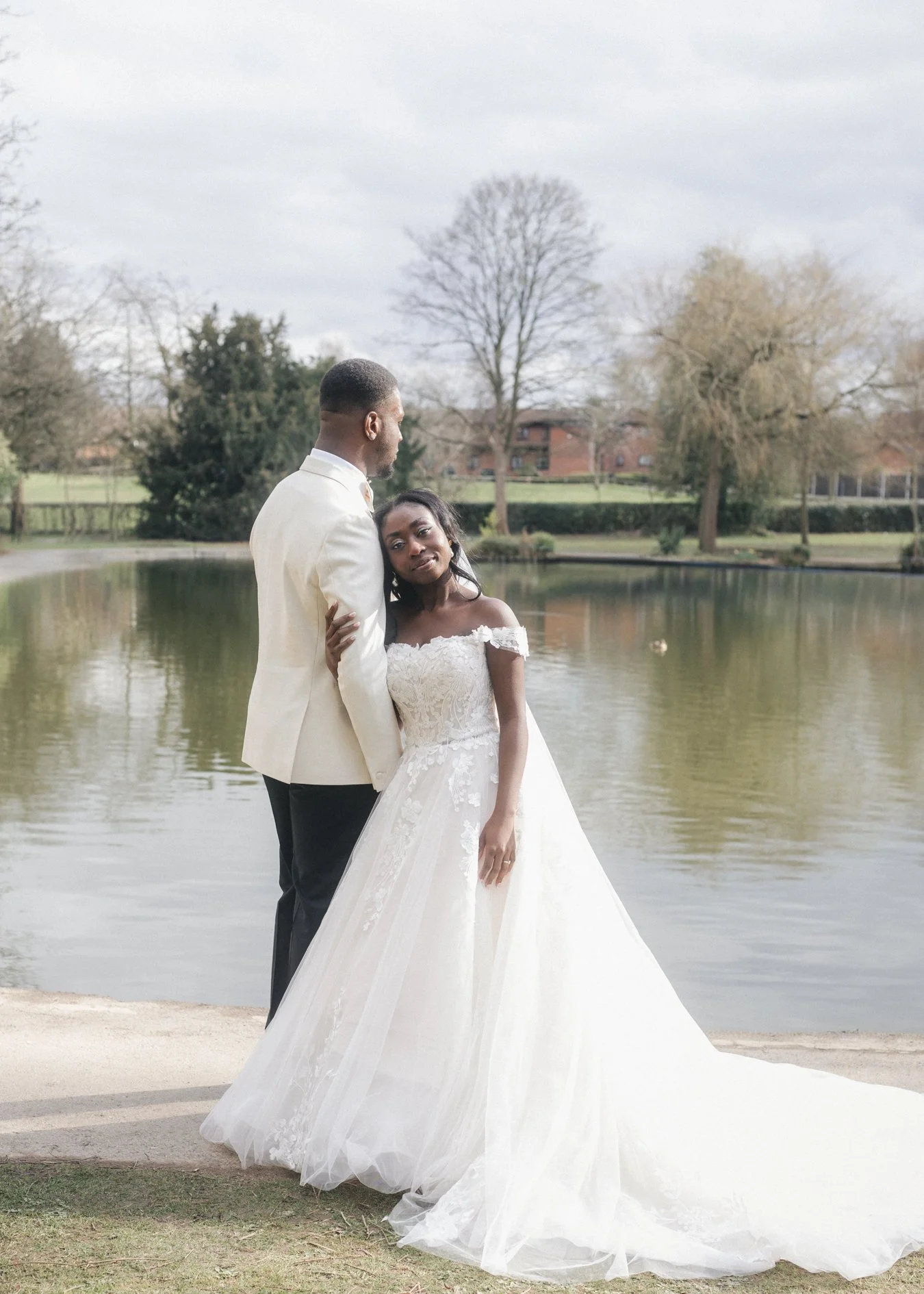 A bride and groom standing by a pond outdoors, with the bride resting her head on the groom's chest, in wedding attire.