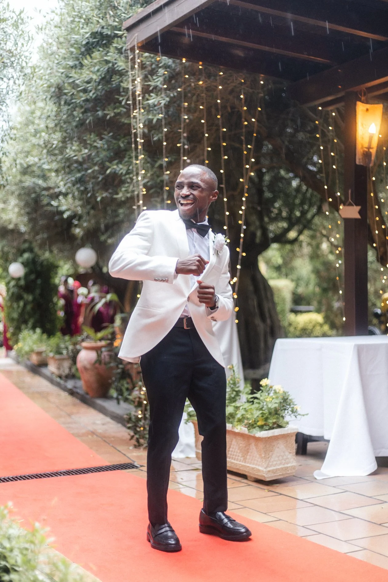 A man in a white tuxedo jacket, black pants, and black shoes dancing outdoors on a red carpet at a wedding or celebration. He is smiling and surrounded by decorative lights and greenery.