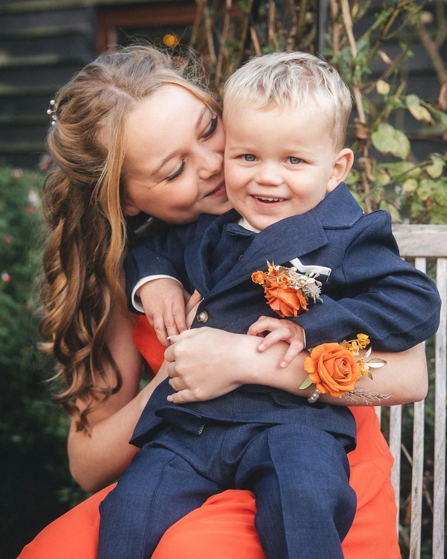 A young woman with long, wavy red hair is holding a smiling little boy in a navy blue suit with orange flowers on his lapel. They are sitting outside in a garden, with the woman kissing the boy on the cheek. The boy is looking at the camera with a bi