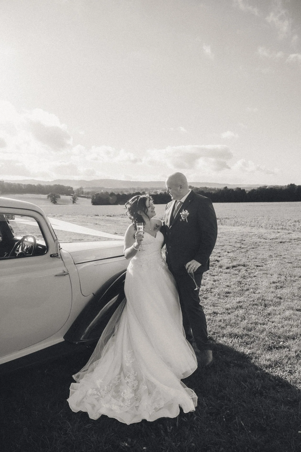 A bride and groom standing outside on a grassy field near a vintage car, holding glasses of champagne and smiling at each other during their wedding celebration.