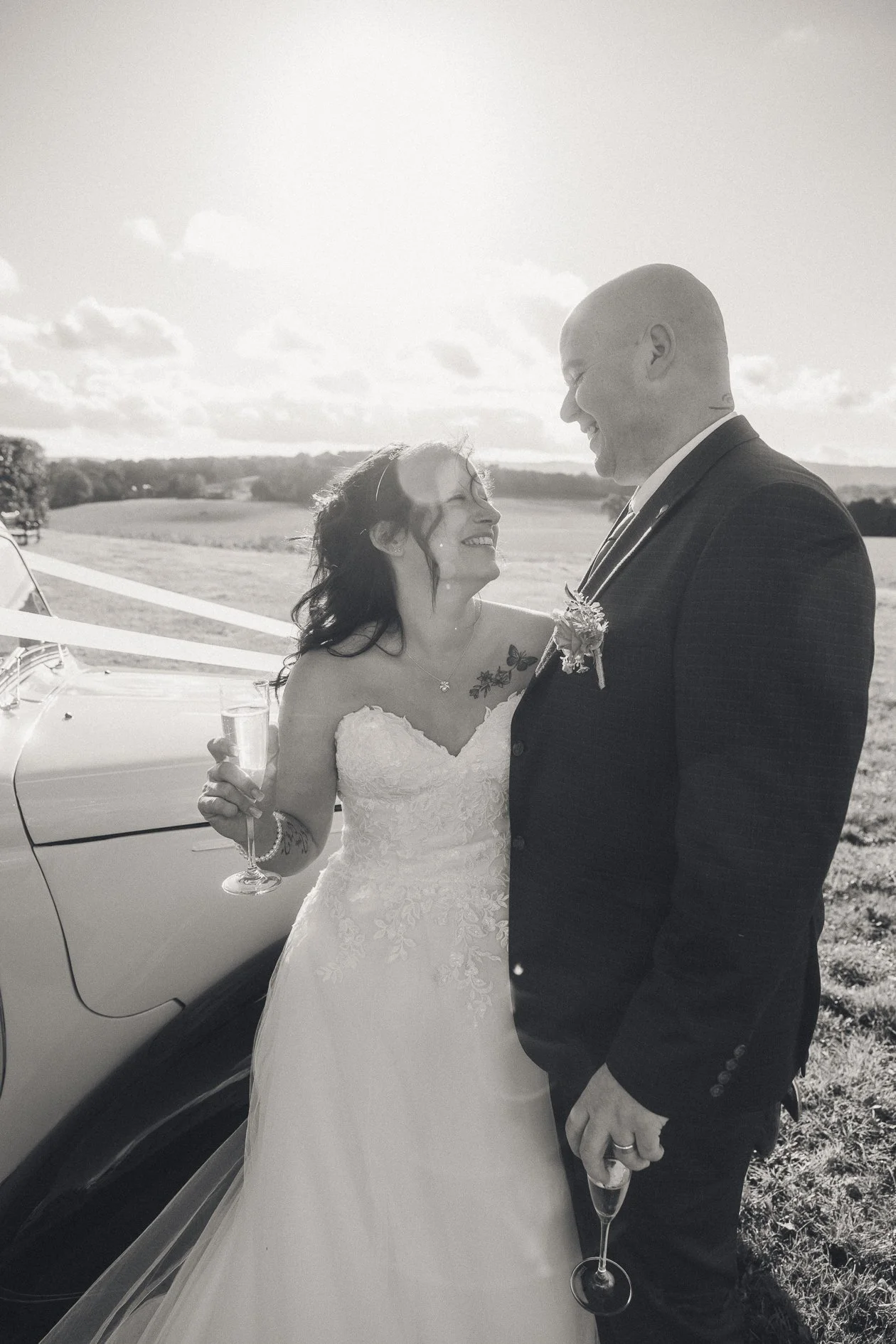 Black and white photo of a bride and groom smiling at each other outdoors during daytime, holding champagne glasses, with a car and open field in the background.