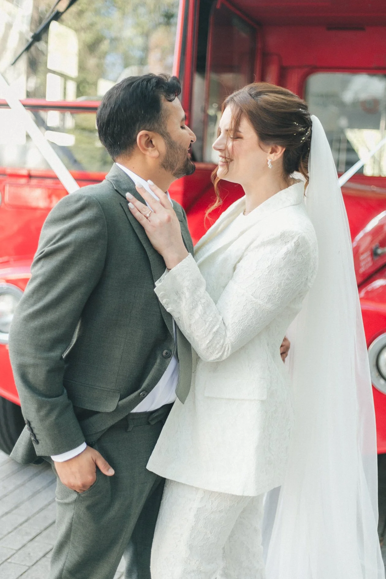 A newlywed couple standing close together in front of a red vintage London bus, smiling with their foreheads touching, dressed in wedding attire. The bride is wearing a white lace suit with a veil, and the groom is in a gray suit with a tie.