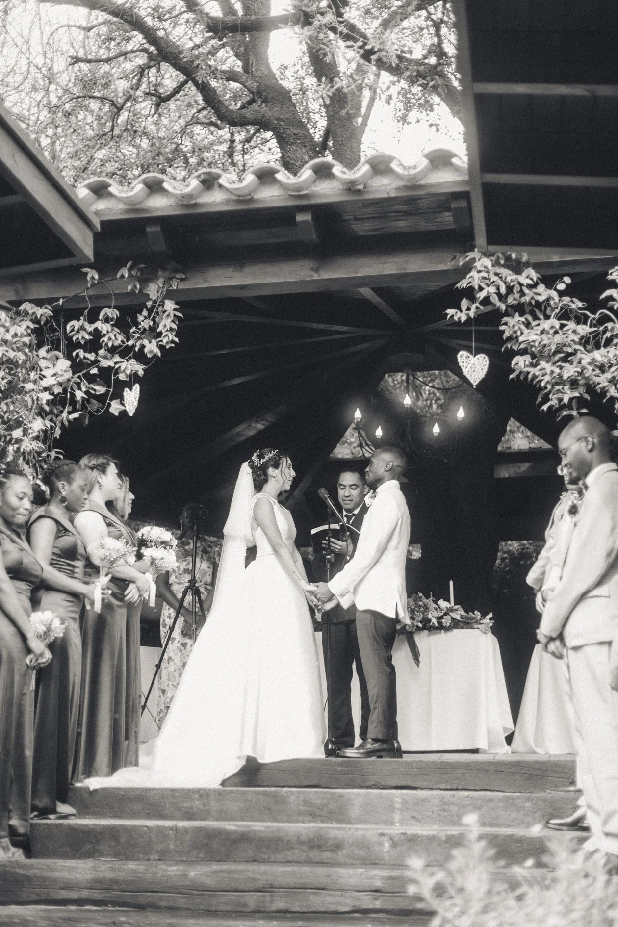A black-and-white photo of a wedding ceremony outdoors, with a bride and groom holding hands and smiling at each other, surrounded by bridesmaids and groomsmen. The setting has a decorated wooden structure with hanging hearts and string lights, and t