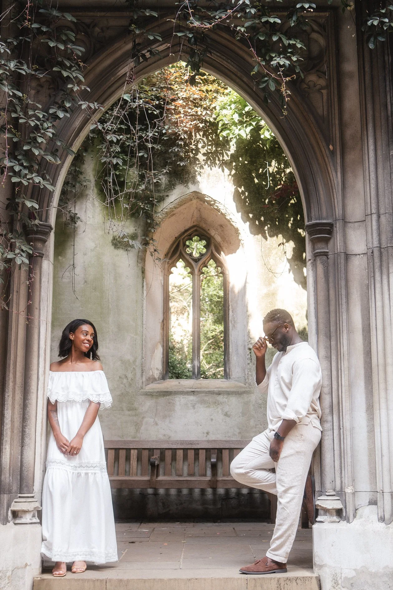 A woman in a white off-the-shoulder dress smiling at a man in beige attire, who is adjusting his sunglasses in a historic, overgrown church ruin with stone columns and a window with a quatrefoil design.