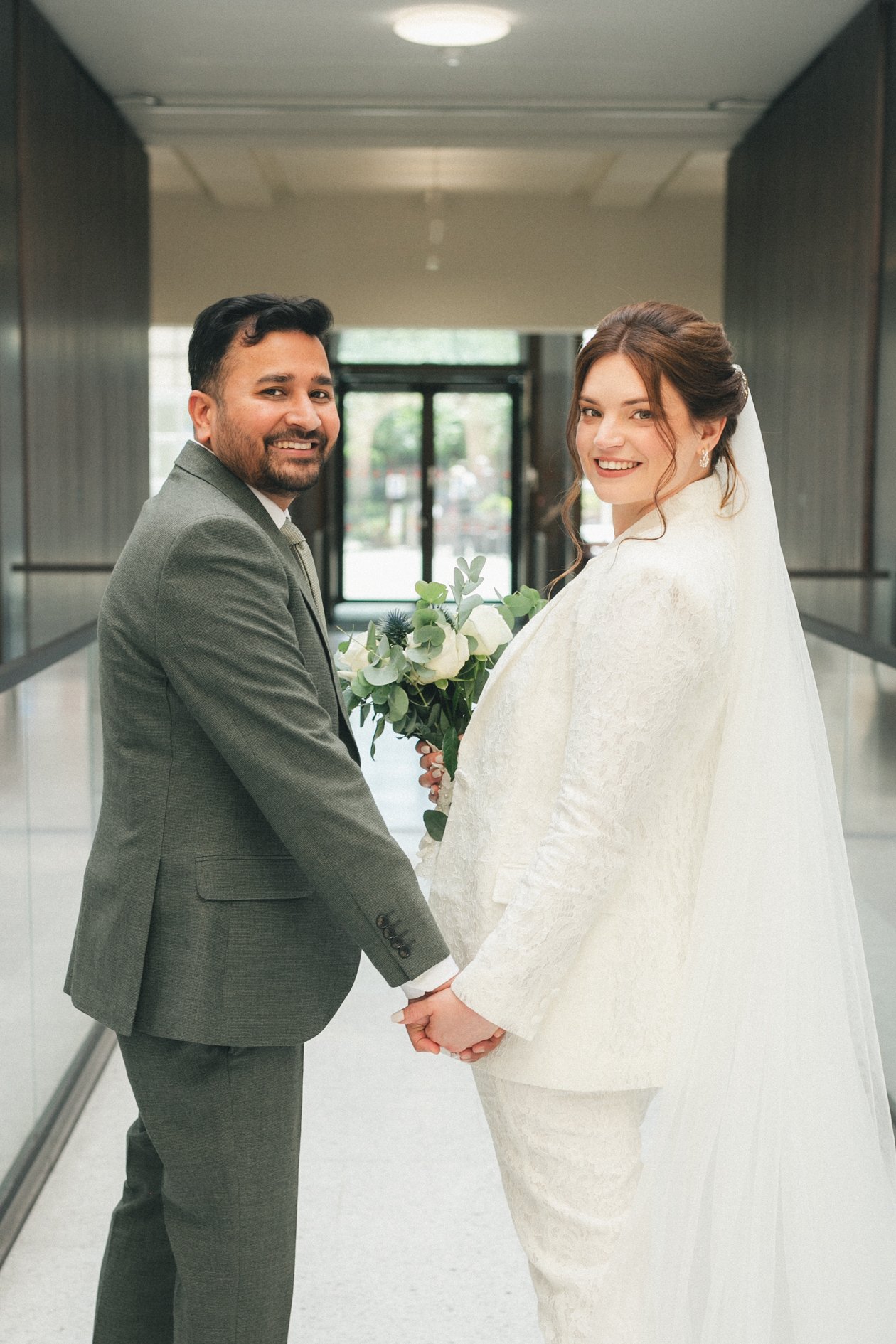A bride and groom holding hands and smiling at each other, standing in an indoor corridor with modern decor, the bride holding a bouquet of white and green flowers.